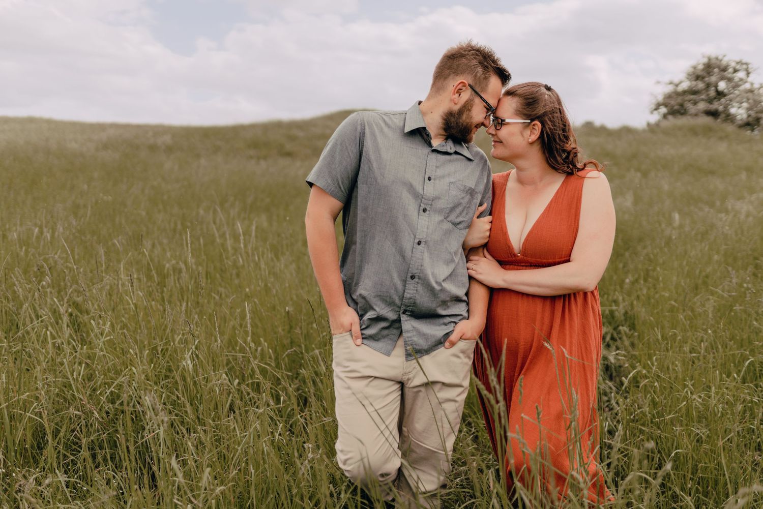 A couple stands lovingly together in a sun-lit grassy meadow, with her wearing a red dress and him in grey casual attire.