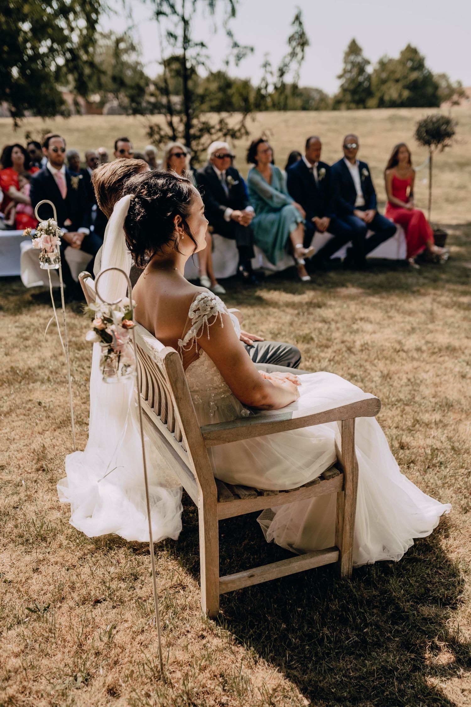 Back view of bride in lace dress seated at outdoor ceremony with guests in background.