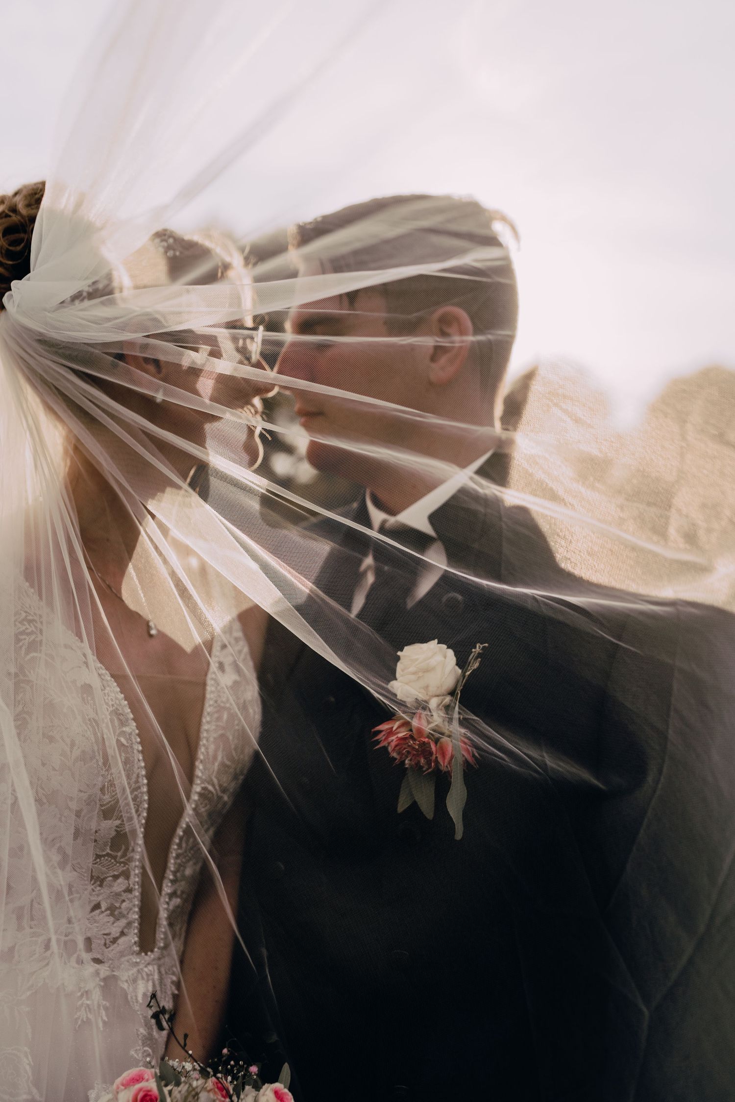 Romantic wedding moment captured through delicate veil with white rose boutonniere detail on black suit.