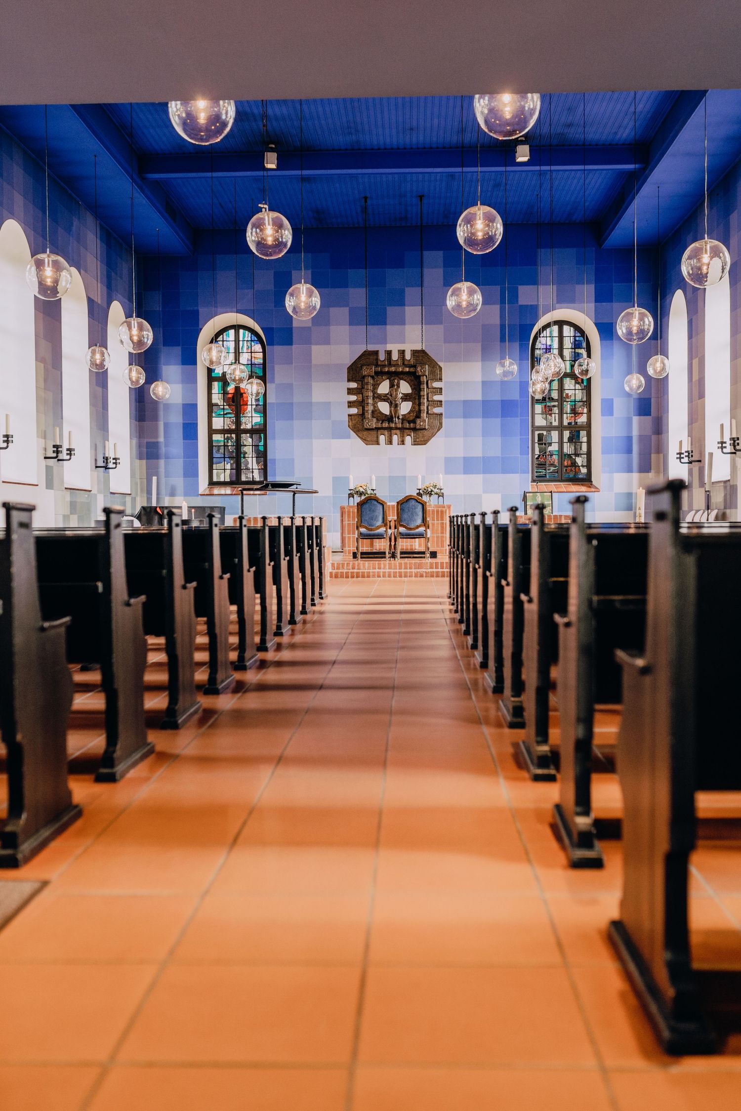 Interior view of a church sanctuary with wooden pews, stained glass windows, and globe pendant lights under a blue ceiling.