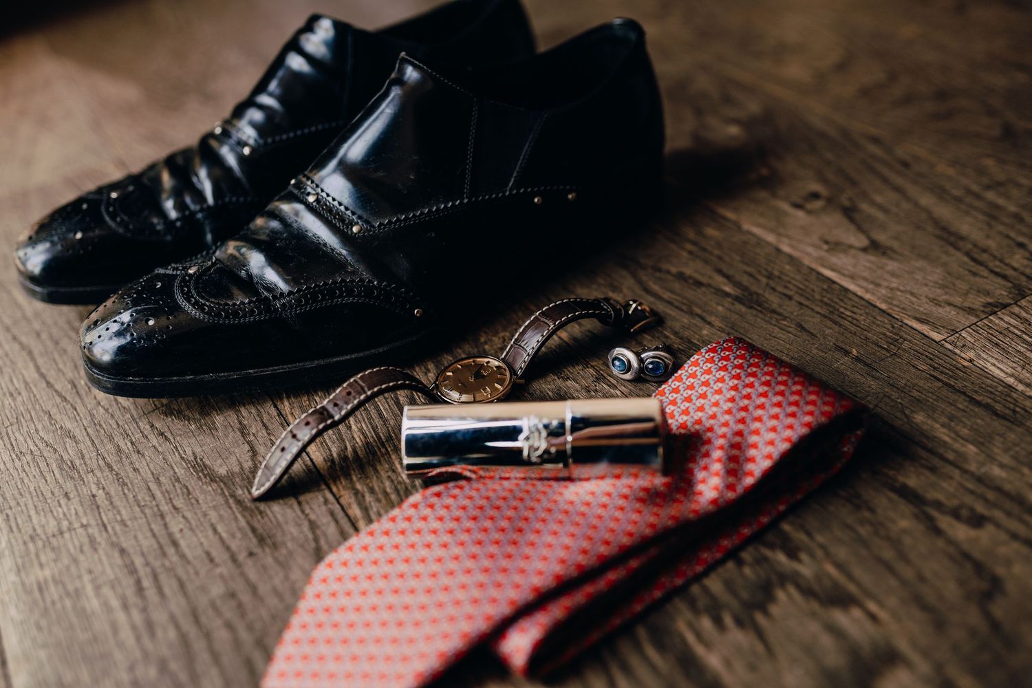 Formal black leather shoes with red tie and accessories on wooden surface.