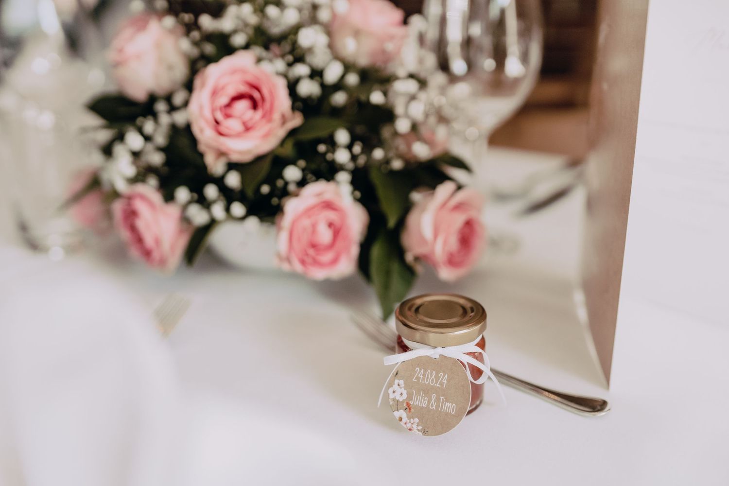 Beautiful pink roses and baby's breath floral arrangement with a jar of honey wedding favor on white table.
