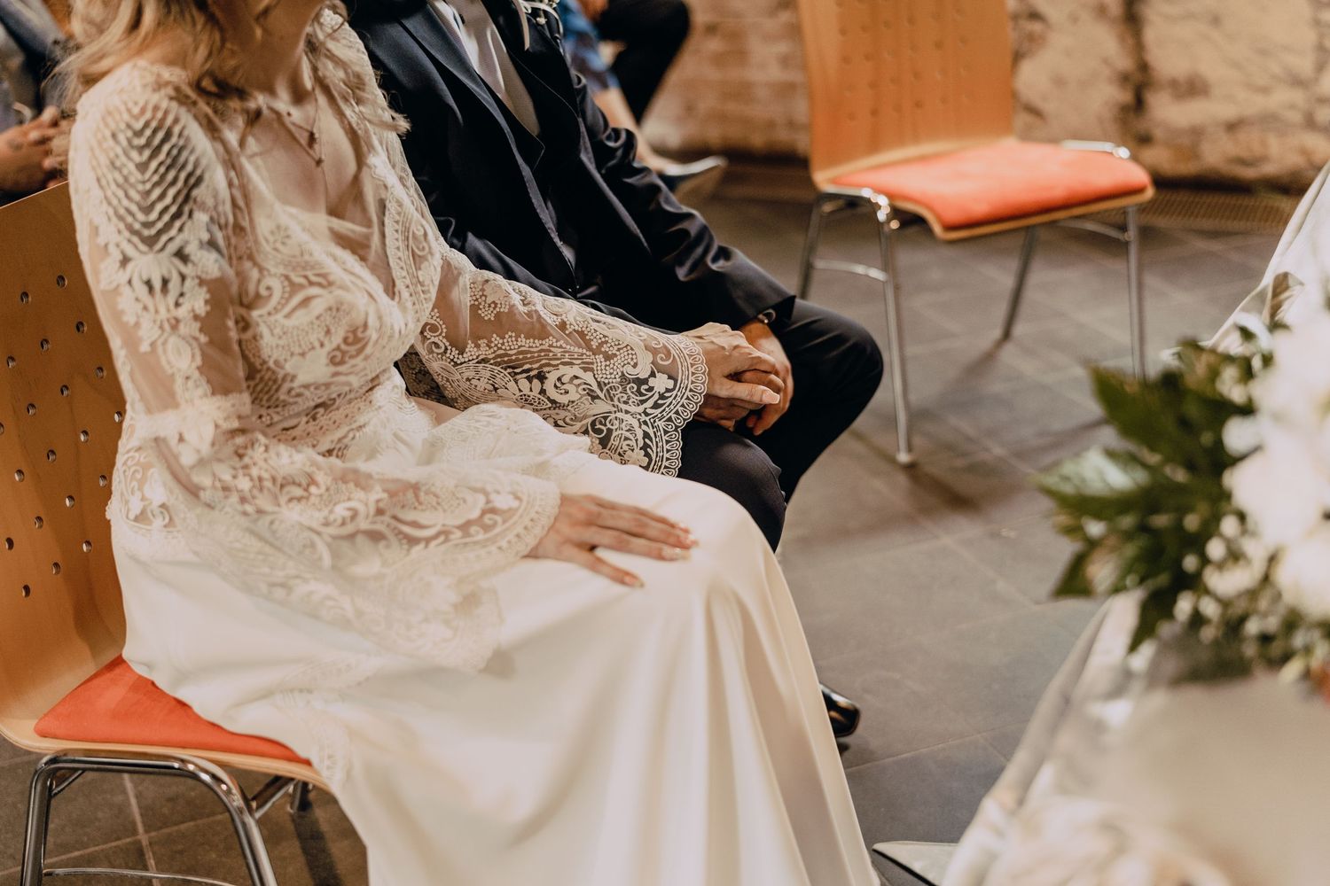 Two wedding guests sit on orange chairs in an indoor venue, with the bride wearing a lace long-sleeved gown.