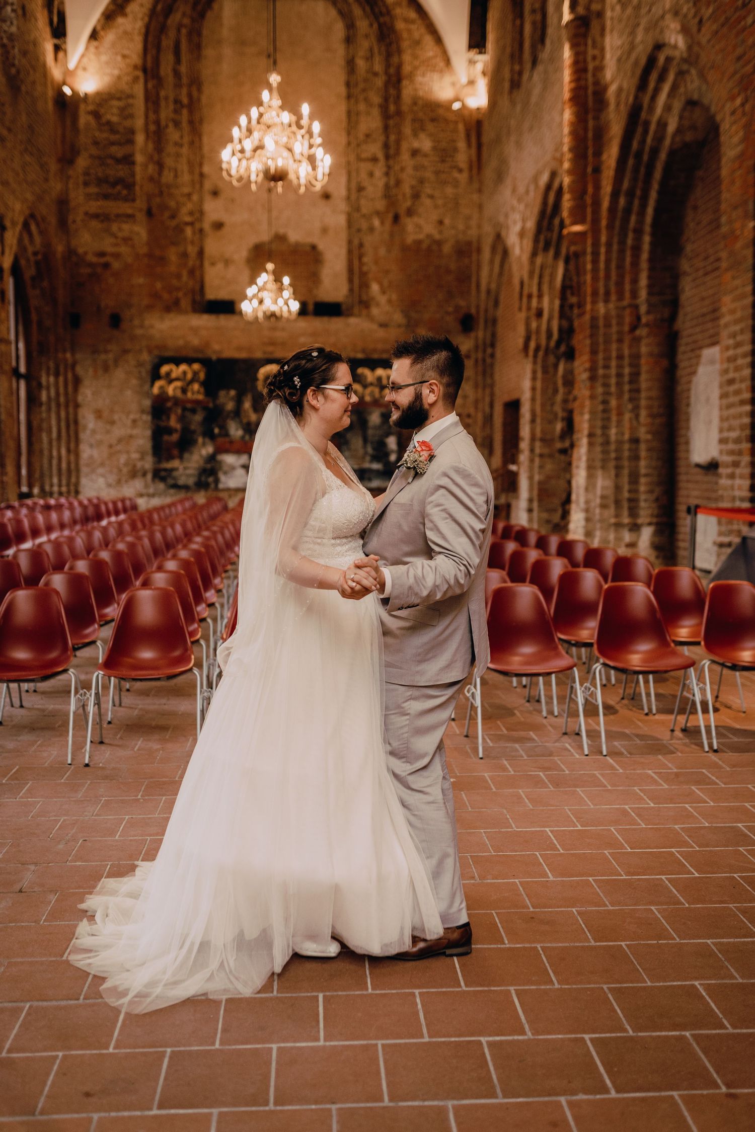 Wedding couple dances in historic brick venue with chandeliers and red chairs.
