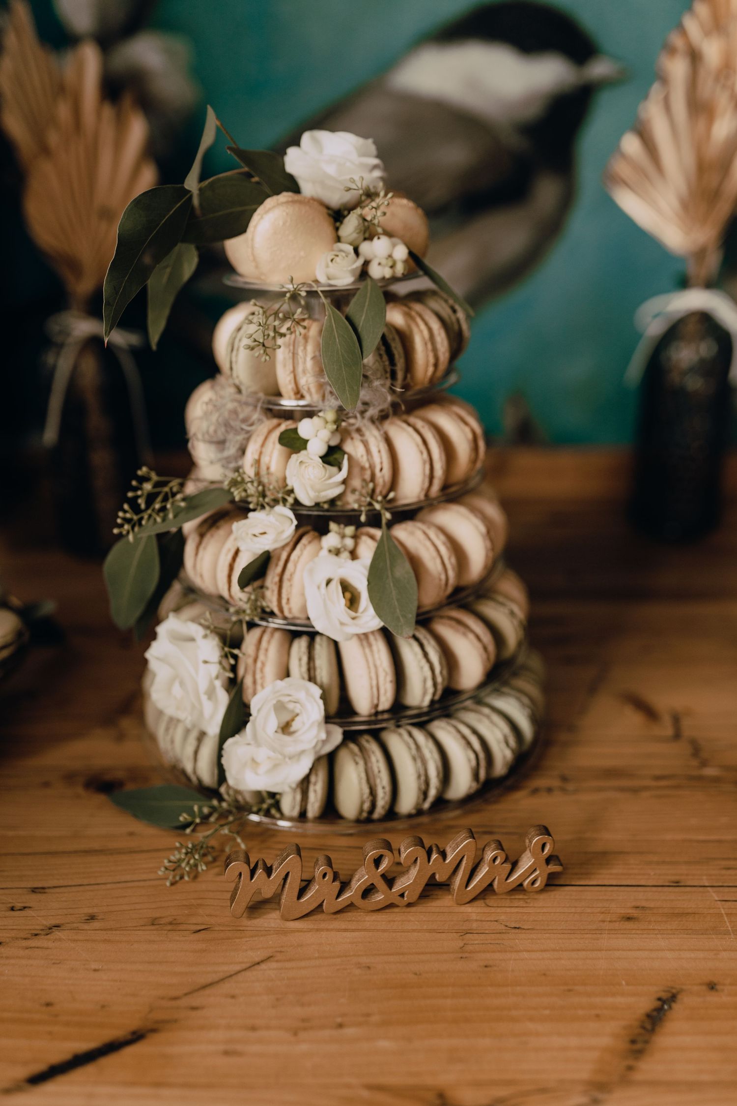 A tiered tower of macarons decorated with white roses and greenery sits on a wooden surface against a teal background.