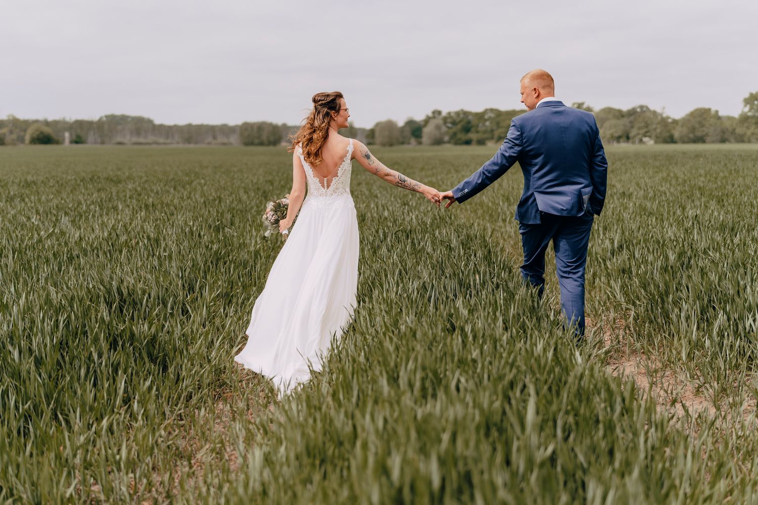 A couple in wedding attire holding hands while walking through a lush green wheat field on their special day.