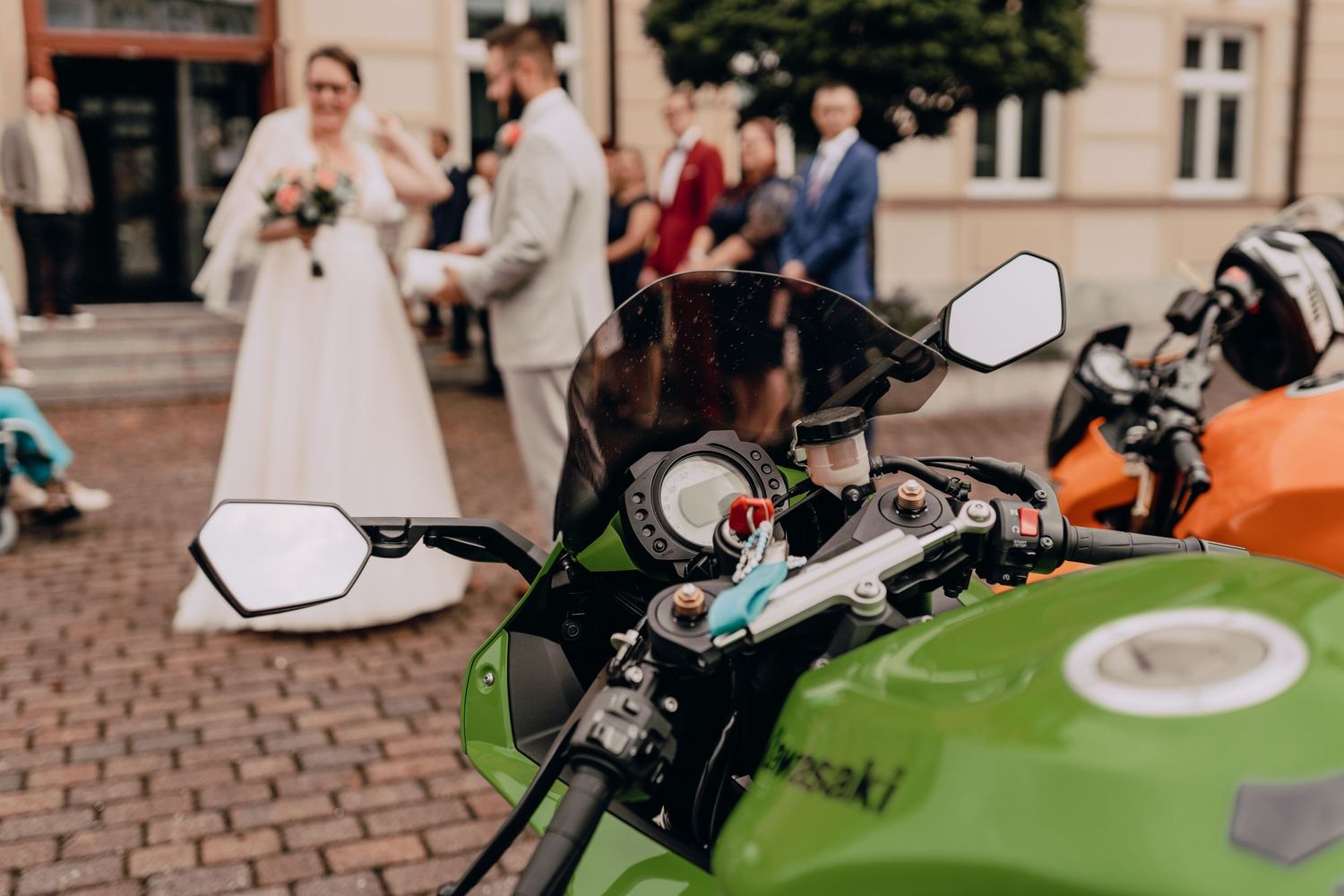 A green motorcycle parked on cobblestone street with a wedding ceremony in the background.