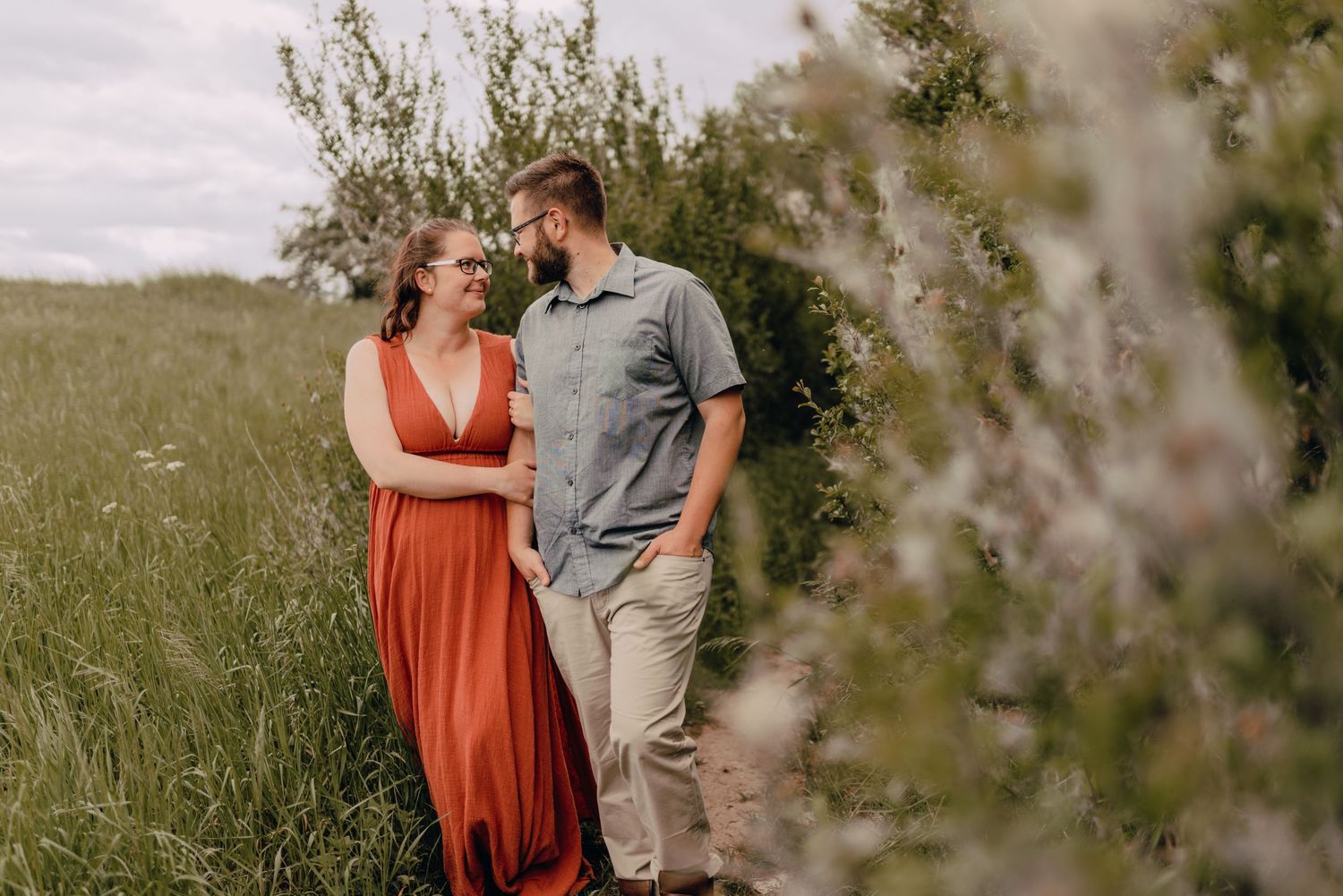 A couple stands together in a scenic field during an outdoor photoshoot with blurred foliage in the foreground.