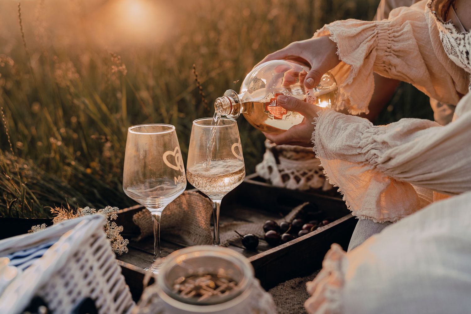 White wine being poured into glasses at a romantic sunset picnic with nuts and snacks on a blanket.