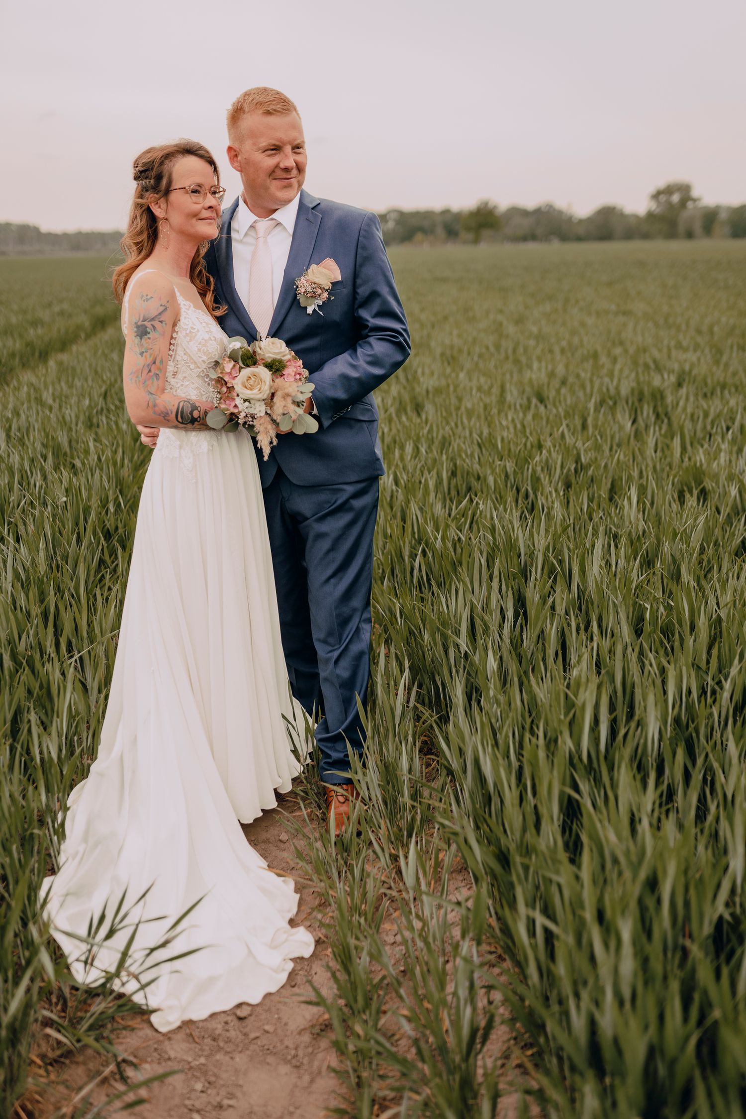 A wedding couple stands together in a green wheat field wearing a white gown and navy suit on their special day.
