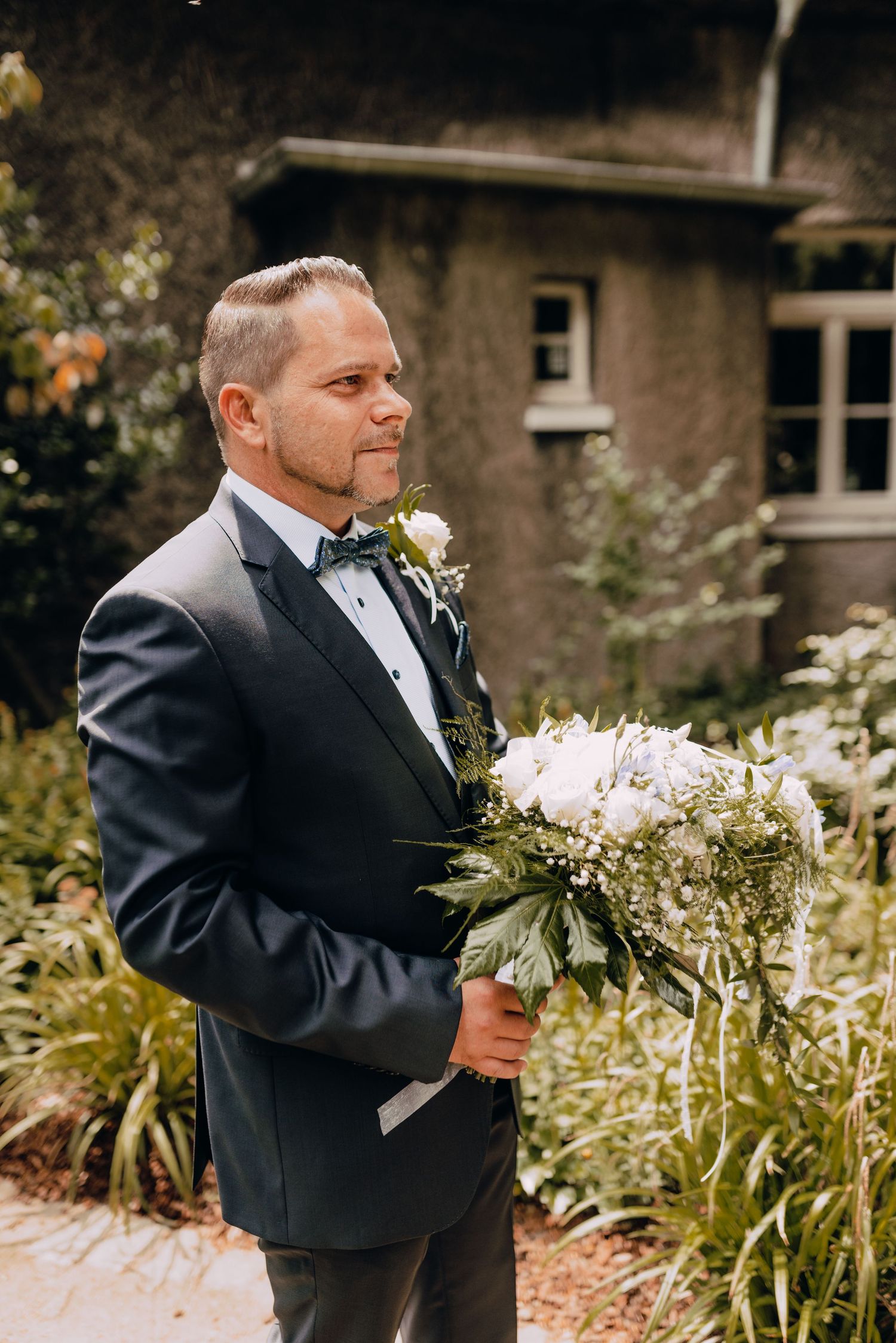 Wedding attire includes classic black tuxedo with bow tie and white boutonniere standing in garden setting.