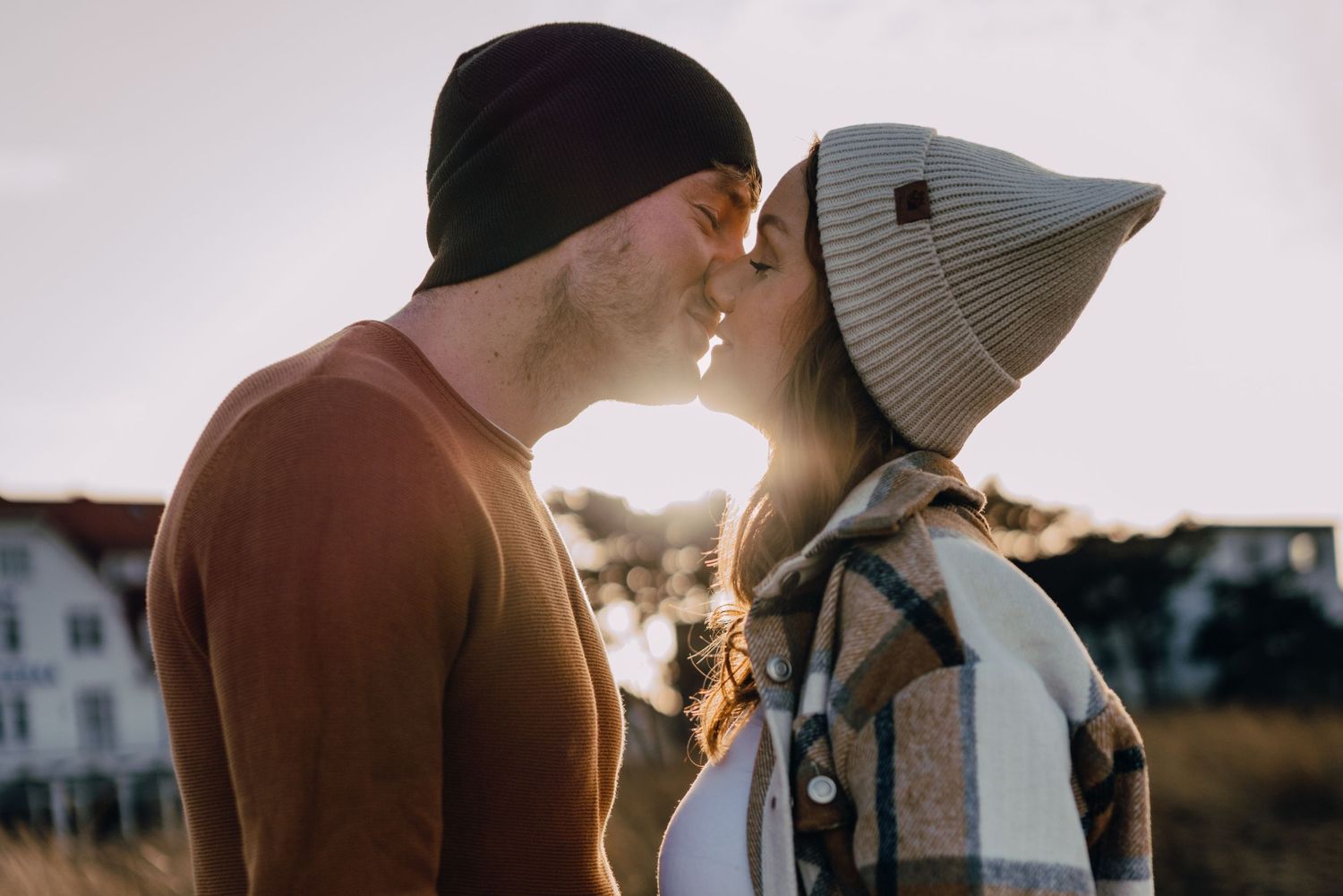 Two people share a tender kiss outdoors while wearing winter hats during golden hour.
