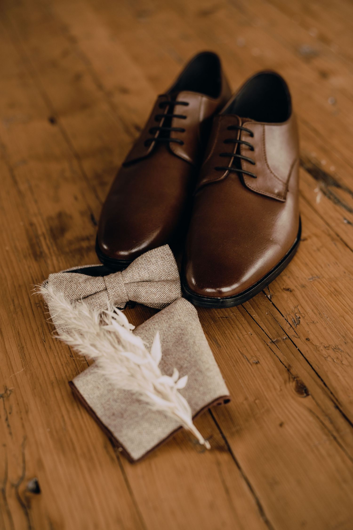 Elegant brown leather dress shoes with beige linen bow tie and white feather on rustic wooden floor.