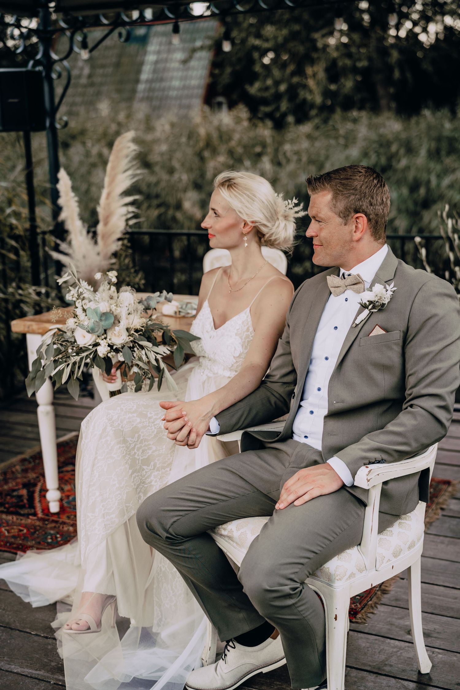 A couple sits together at their wedding ceremony featuring boho decor with pampas grass and outdoor garden setting.