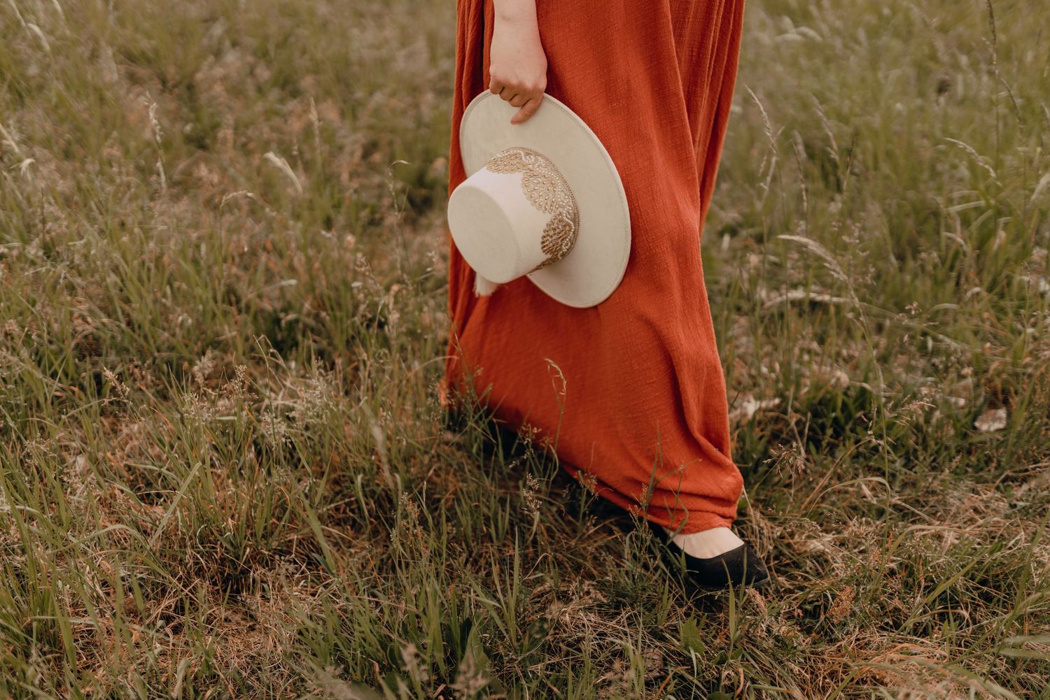 A white sun hat dangles against a flowing red dress in a grassy field.