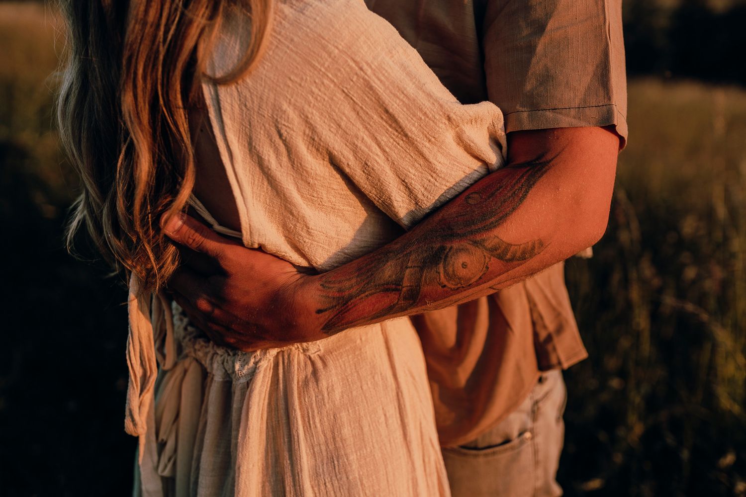 Two people in casual attire hold each other close while standing in a sun-drenched countryside setting.