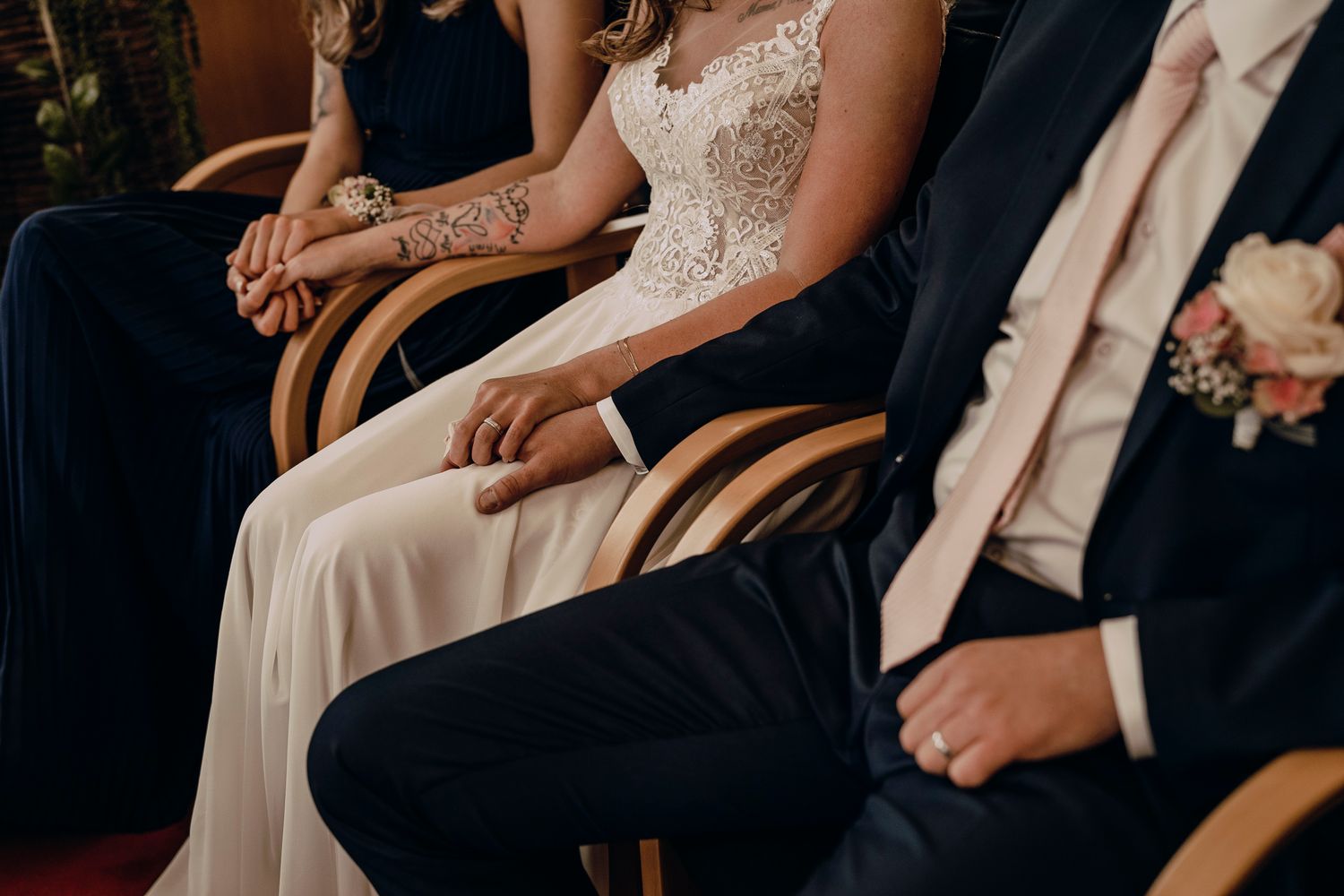 Groom holds his bride's hand tenderly on ornate wooden chairs during an elegant wedding ceremony.