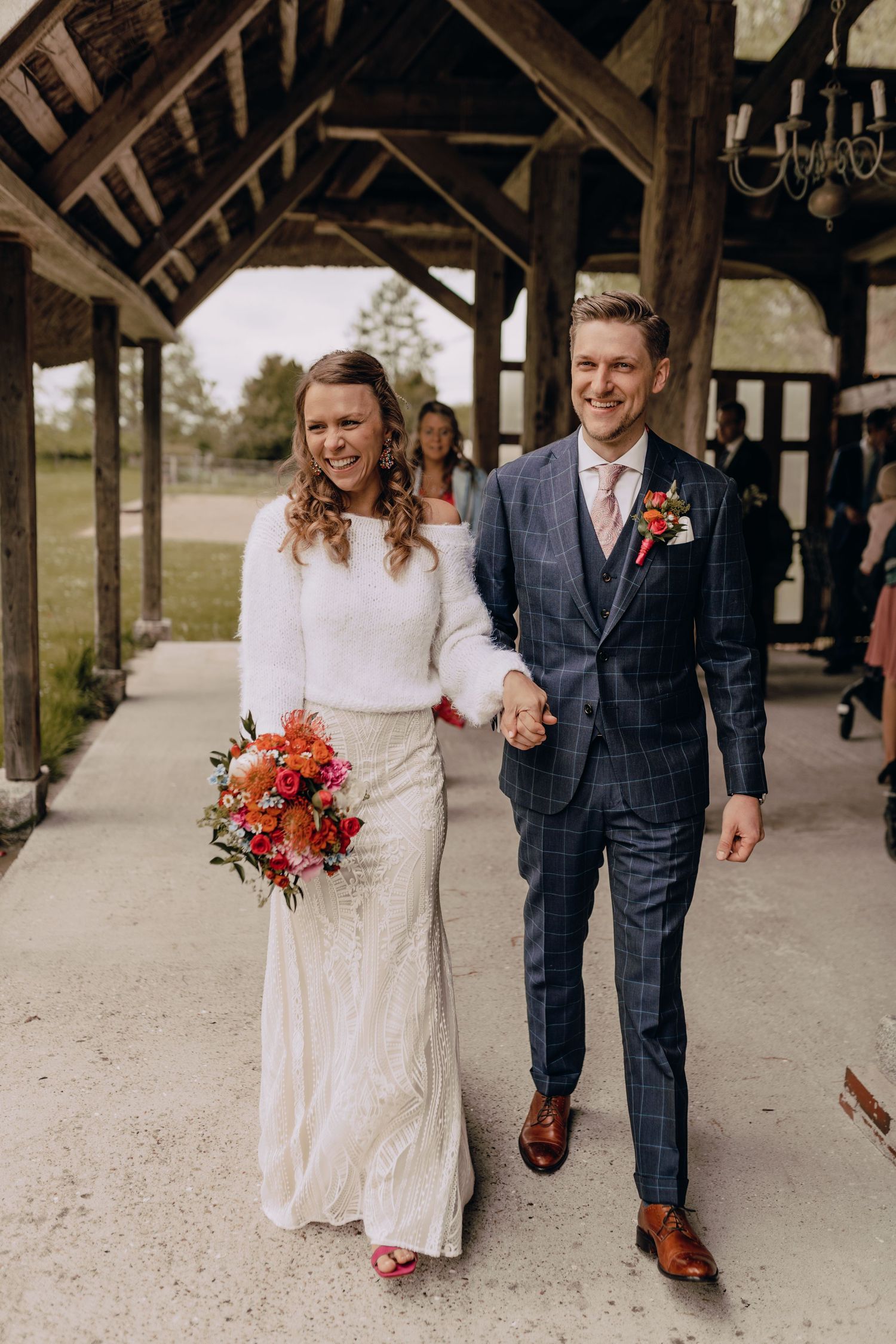 Wedding couple walks under rustic wooden covered walkway with bride holding vibrant autumn bouquet.