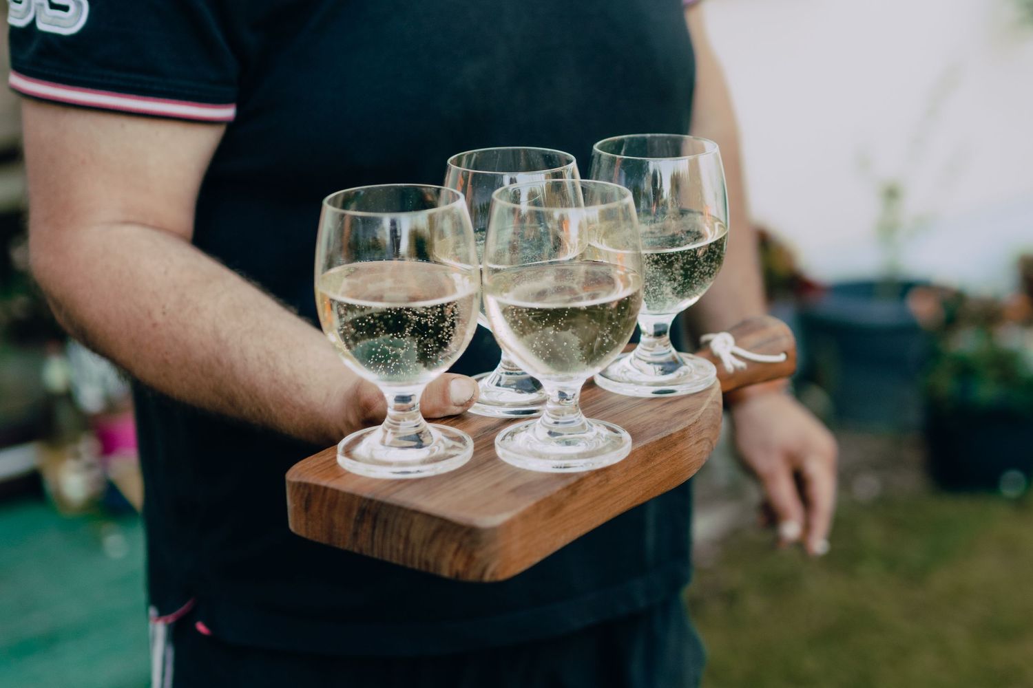 Server holds a wooden tray with five stemmed wine glasses filled with white wine at an outdoor event.