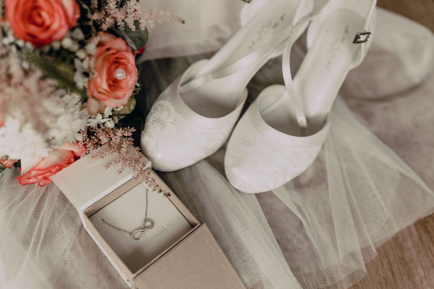 Wedding detail photo showing white bridal shoes, coral roses, and a necklace jewelry box on tulle fabric.