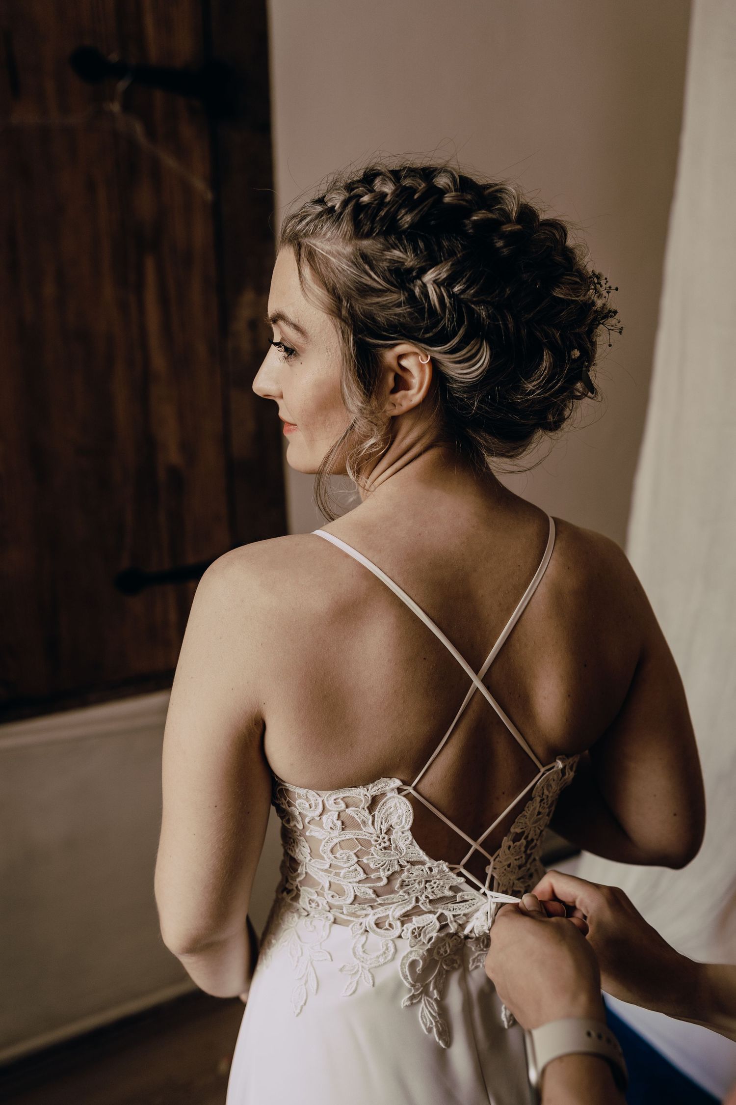 An elegant bride in a lace-backed wedding dress with cross-straps gets help with fastening her gown in a moody setting.
