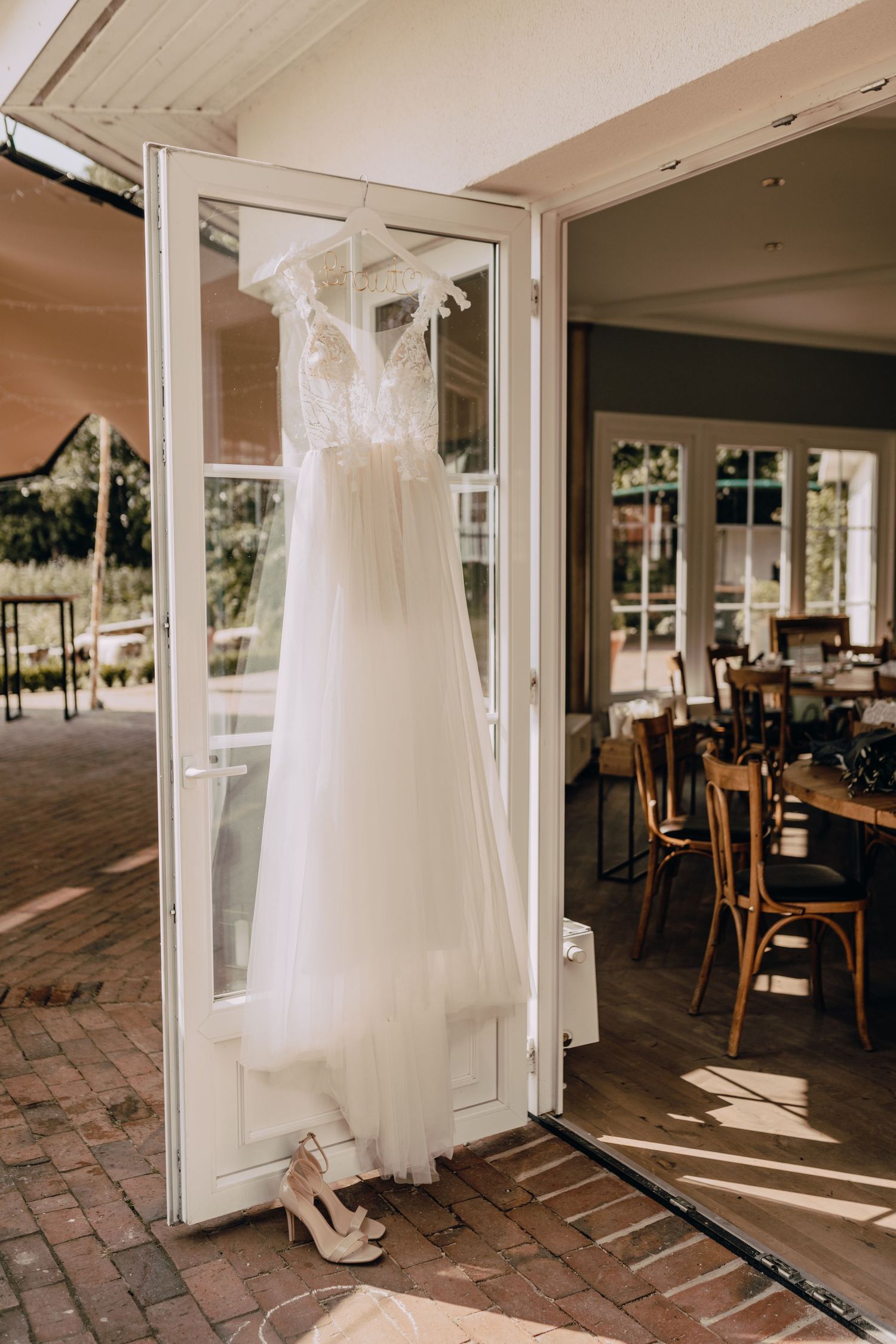 A delicate white wedding dress hangs elegantly in a glass display case on a covered porch with restaurant seating visible.