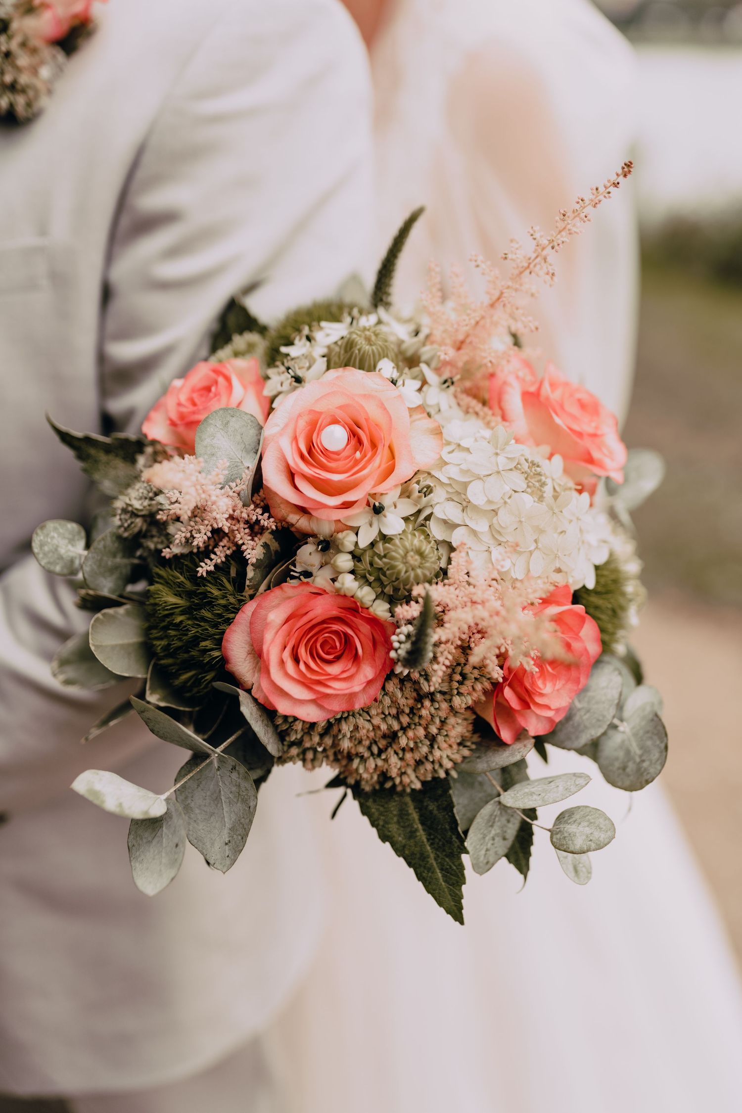 Wedding bouquet with coral roses, baby's breath, and eucalyptus leaves held against white formal attire.