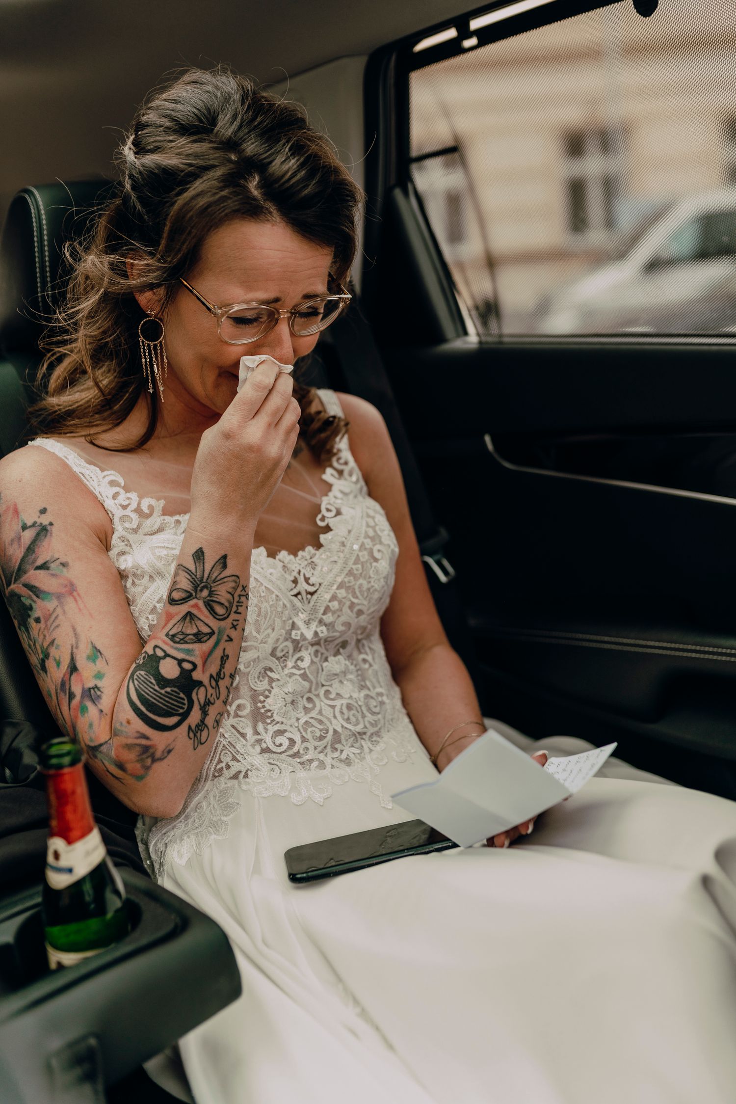 An emotional bride in a lace wedding dress with arm tattoos sits in a car reading something while wiping her eyes.