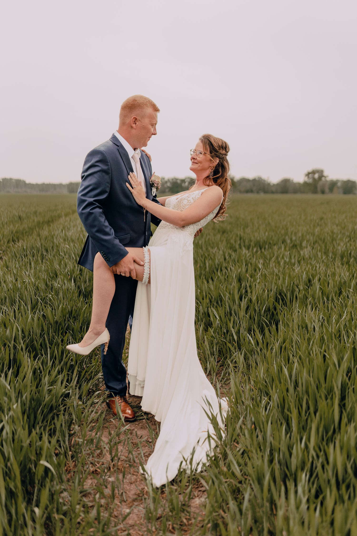 A couple in wedding attire embrace while standing in a lush green wheat field on their special day.