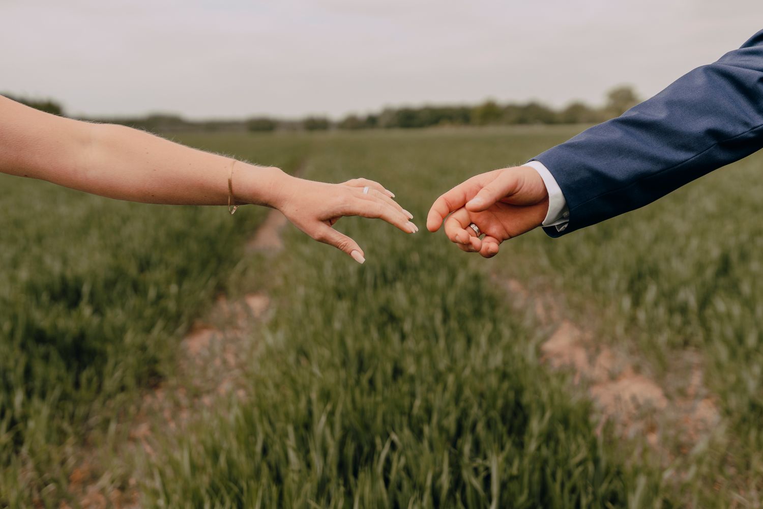 Two people hold hands reaching towards each other in a green grassy field at sunset.
