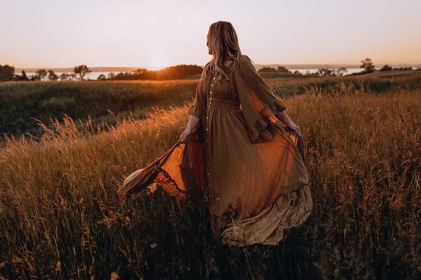 Figure in flowing dress stands in golden field at sunset casting long shadows across tall grass landscape.