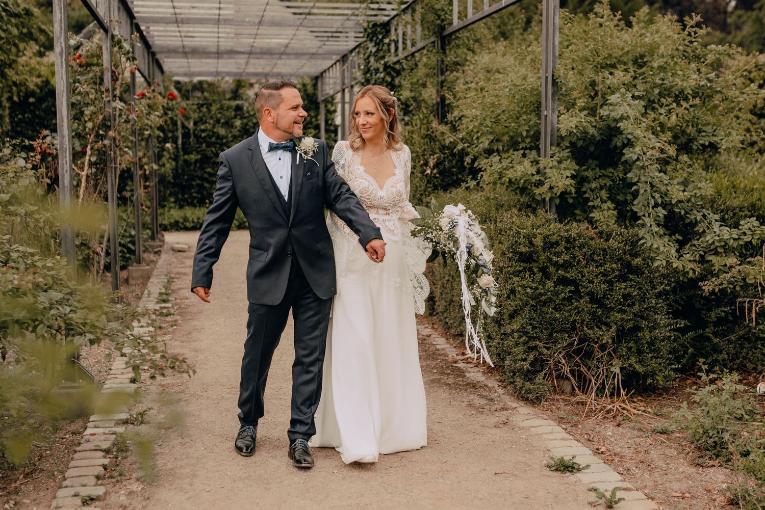 A newlywed couple walks hand in hand through a lush greenhouse pathway on their wedding day.