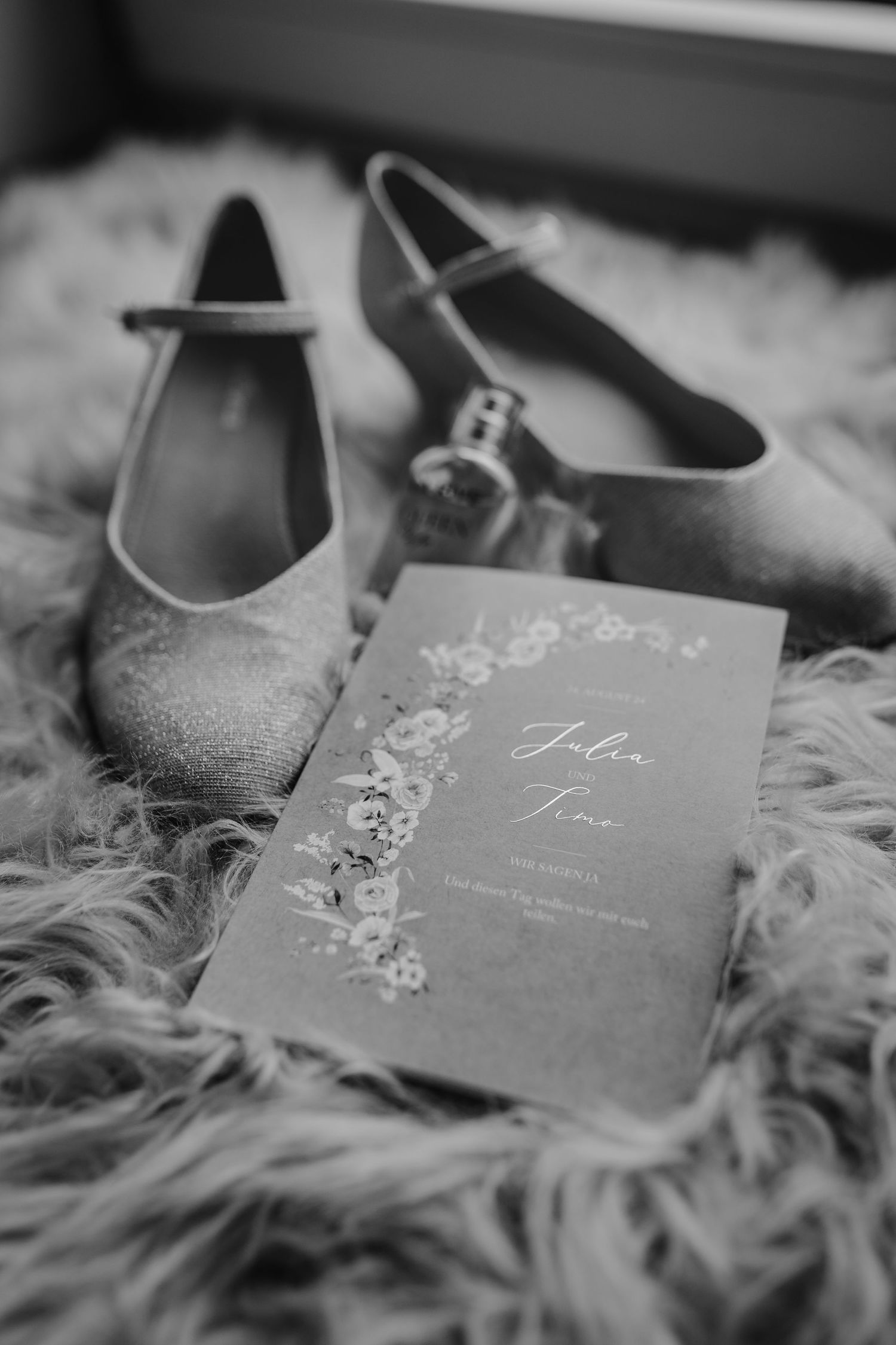 Wedding shoes and invitation card with floral design arranged on soft fur backdrop in black and white.
