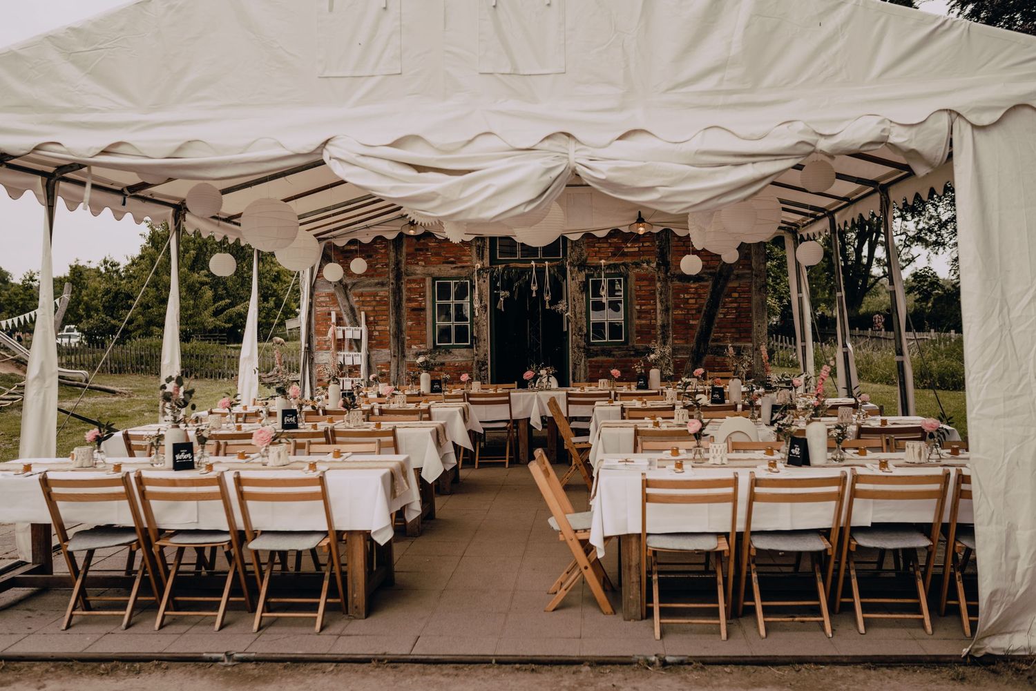 Rustic wedding reception setup under a white tent with wooden chairs and long dining tables.