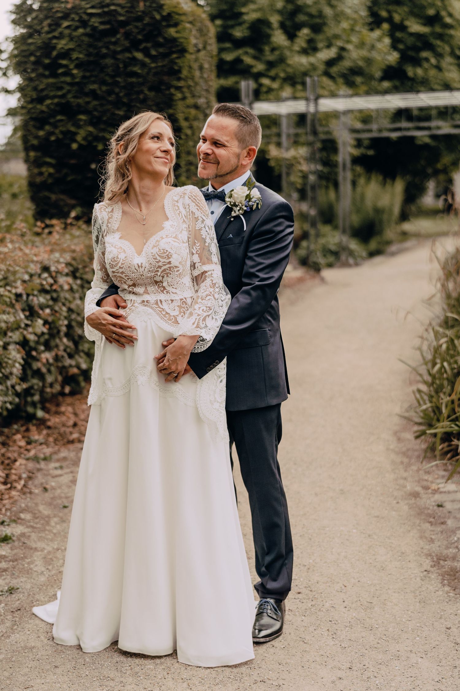 A bride in a lace detailed white gown and groom in navy suit embrace on a garden path during outdoor wedding.