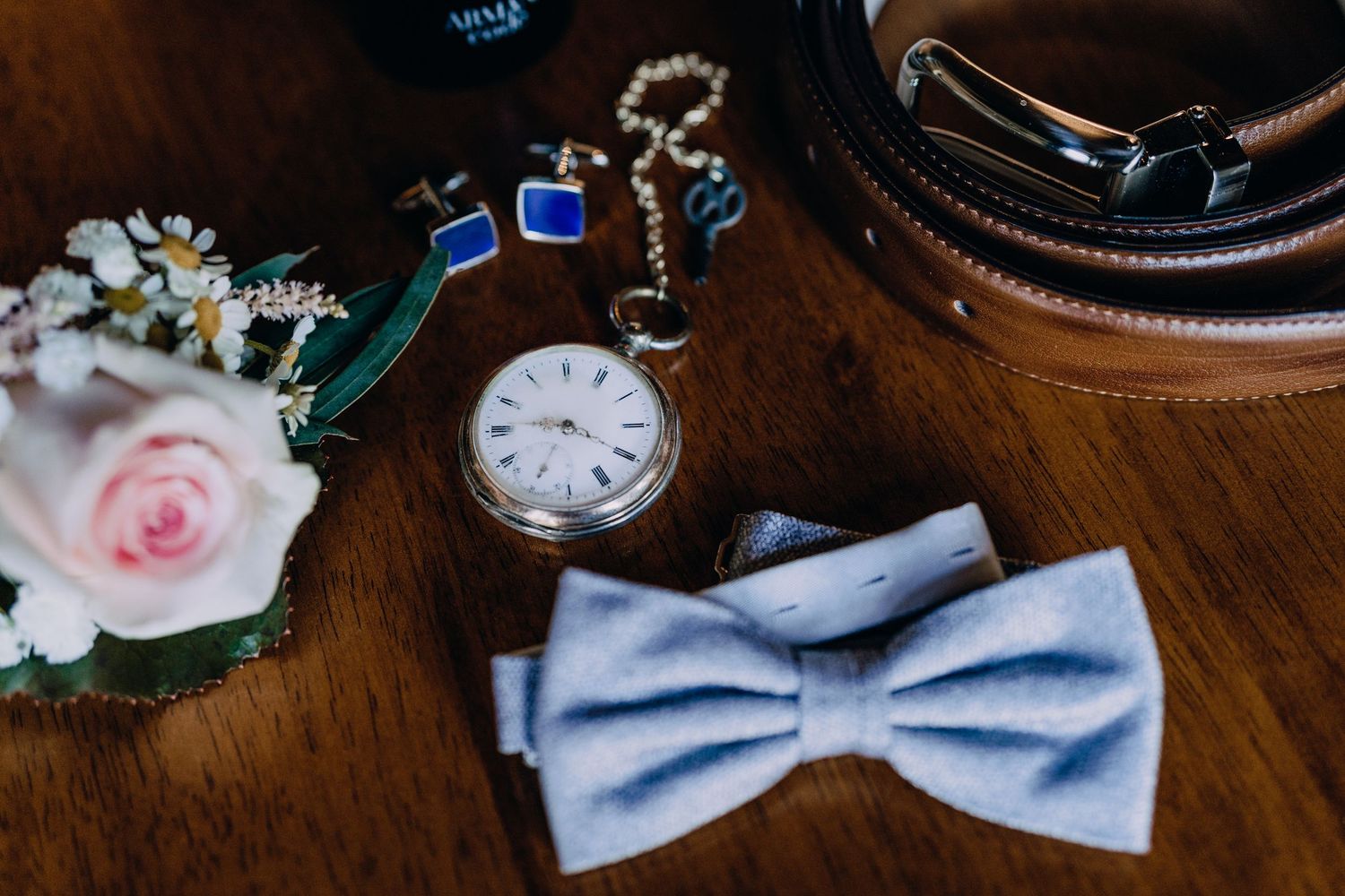 Wedding accessories including a light blue bowtie, pocket watch, and pink rose arranged on wooden surface.