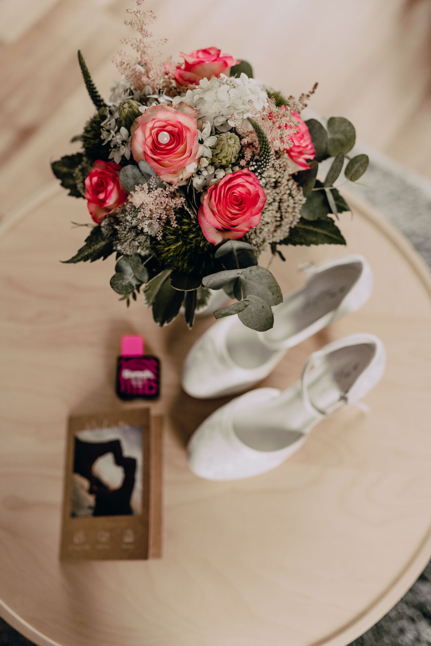 Pink and white rose bouquet with eucalyptus leaves next to white bridal shoes on round table.