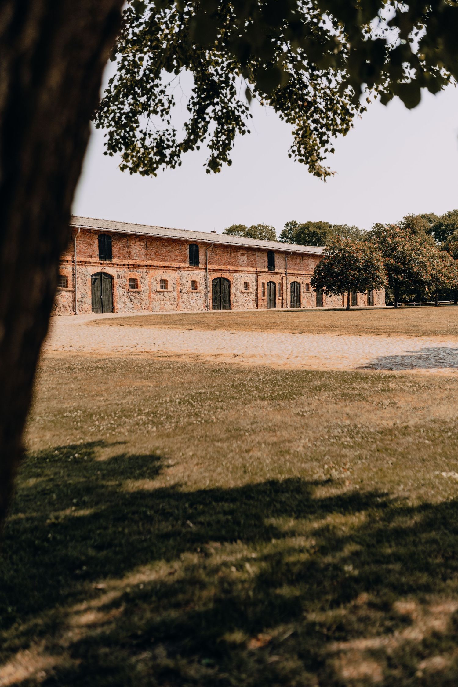 Long red brick stable building with white trim sits on a grassy hillside under a partly cloudy sky.