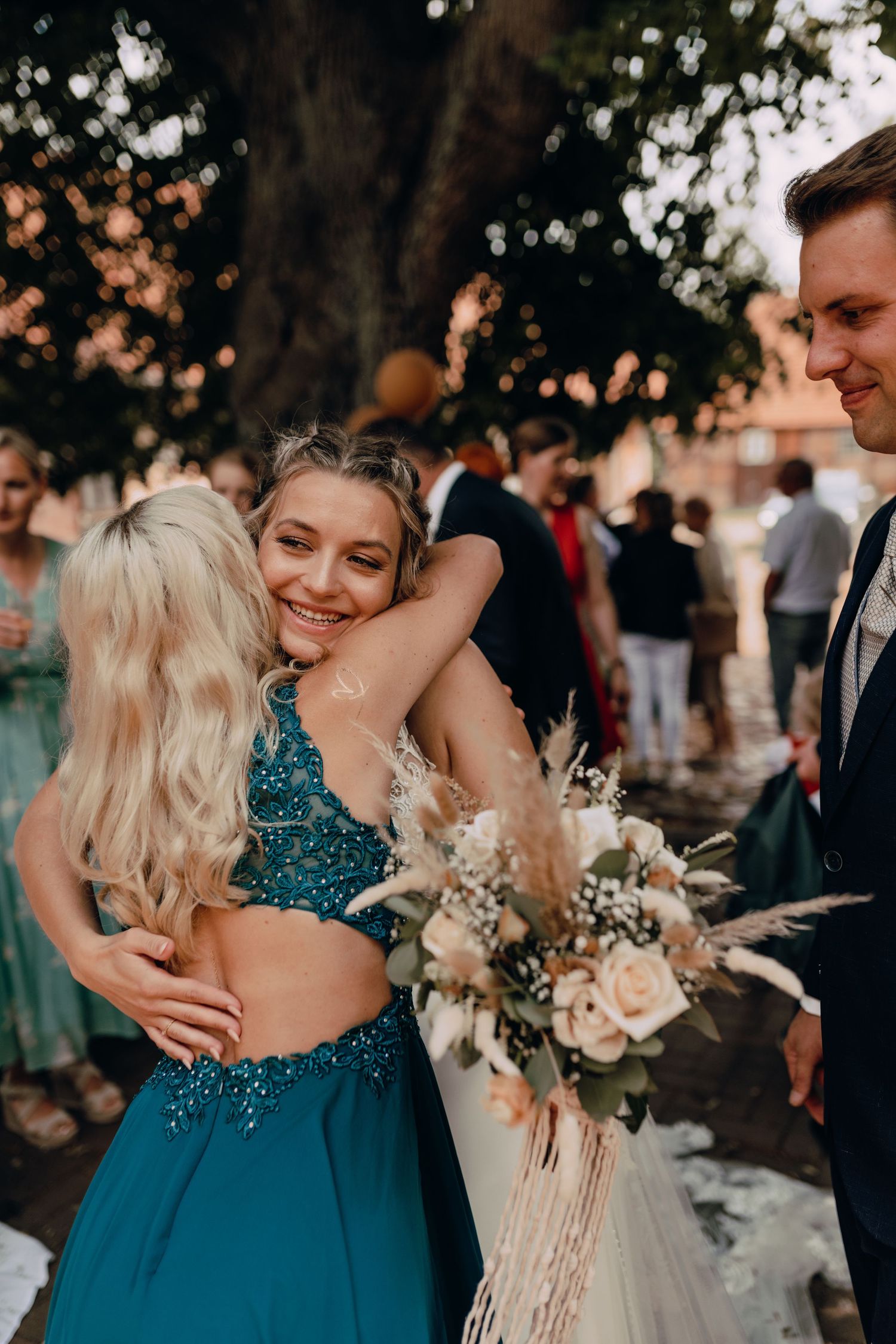 Two people embrace while another holds a bouquet at an outdoor wedding gathering with teal dresses and formal attire.