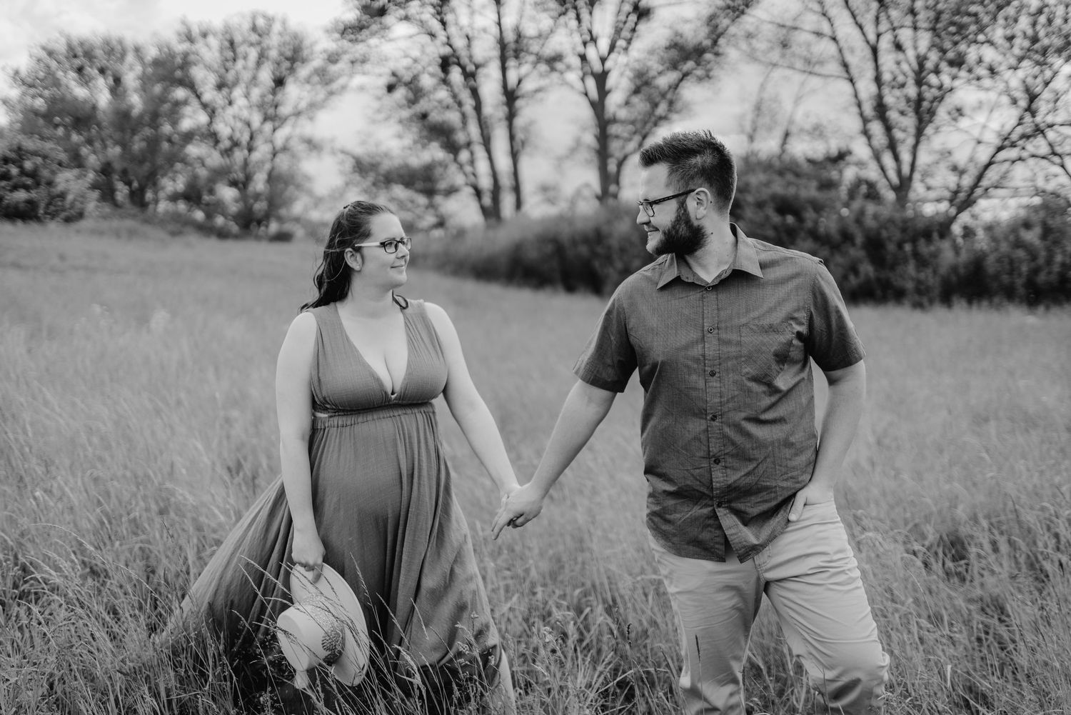A couple laughs and walks together through a grassy meadow in summer during their outdoor photoshoot.