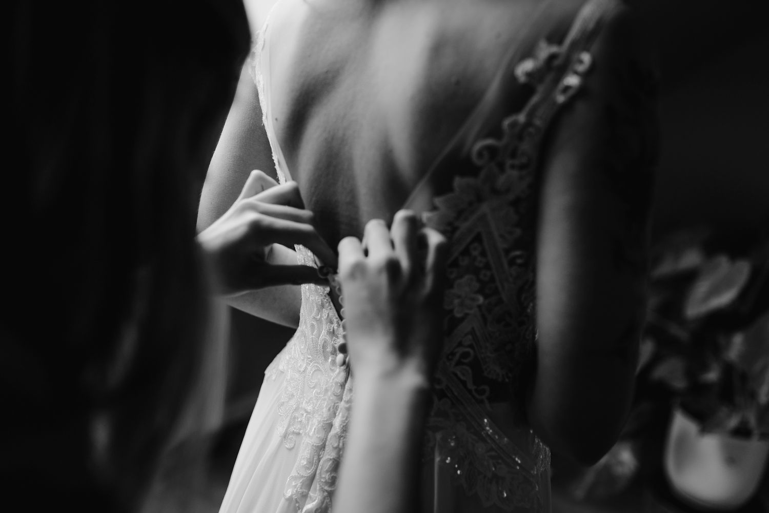 Hands helping to fasten buttons on a delicate white lace dress in black and white photography.
