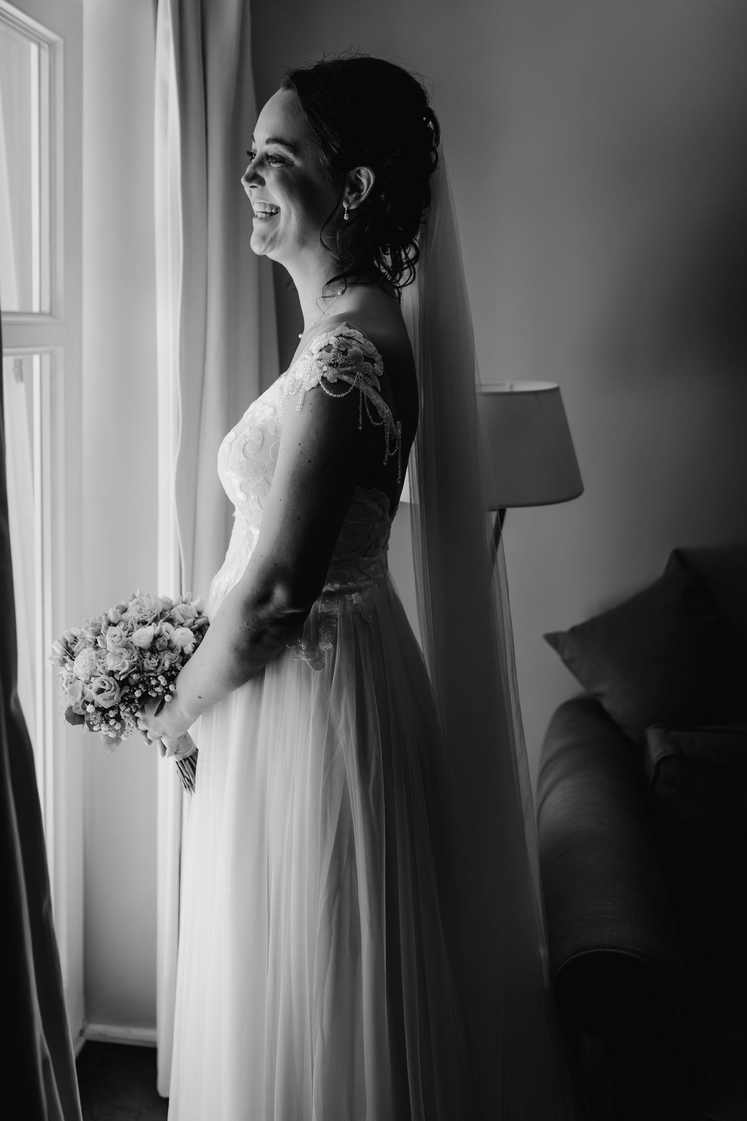Bride in elegant lace gown stands by window holding bouquet in black and white portrait.