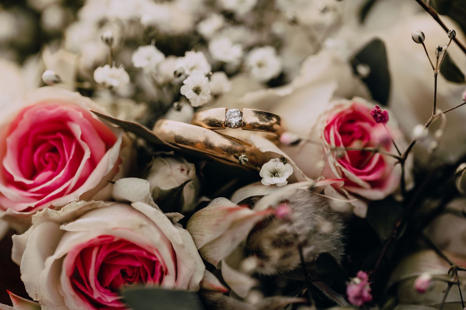 Gold wedding rings nestled among pink roses and white baby's breath flowers in a romantic floral arrangement.