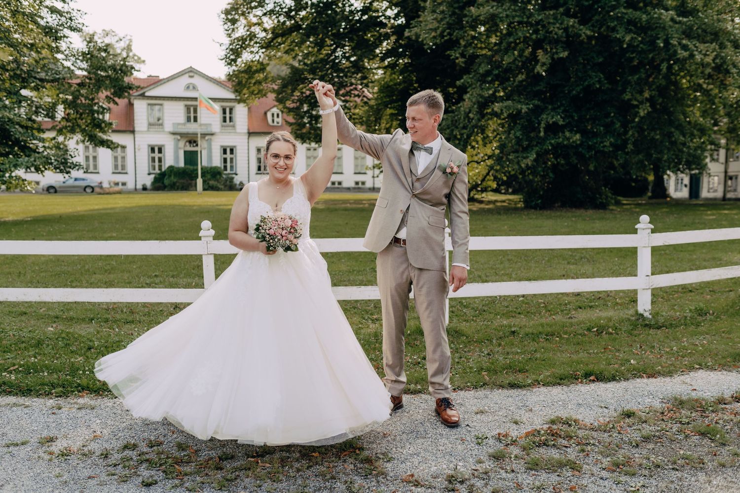 A couple pose together in formal wedding attire on a gravel path with a white picket fence and house in the background.