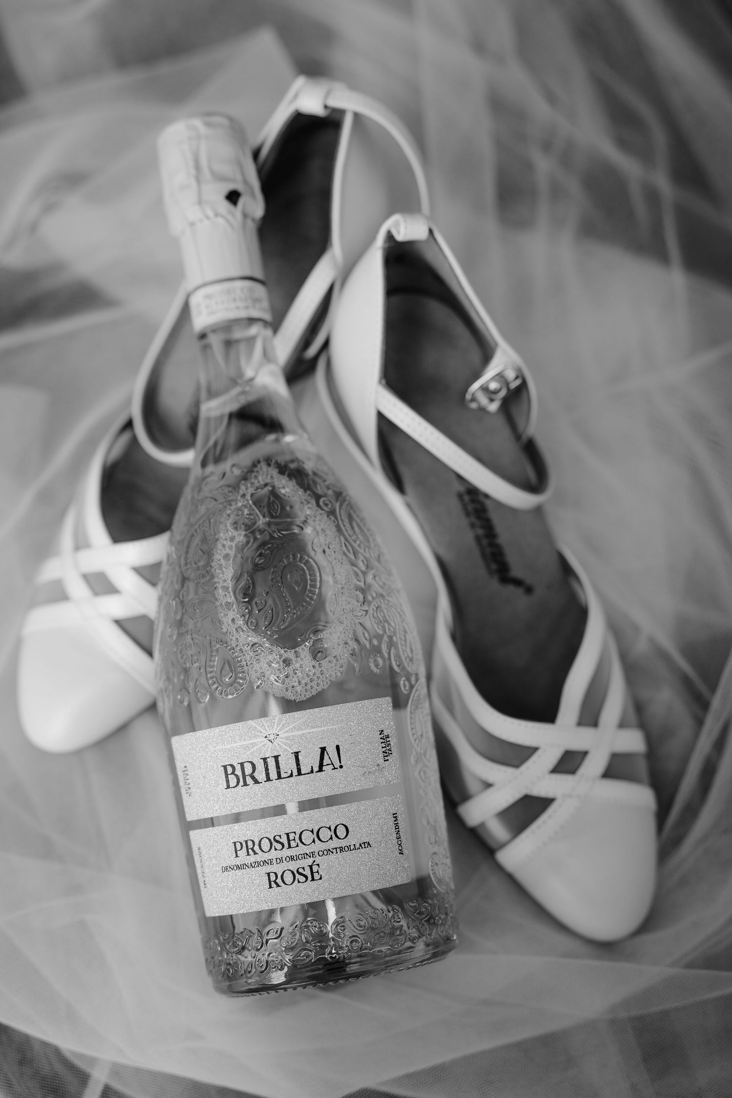 A bottle of Brilla Prosecco Rose positioned next to elegant white bridal shoes on tulle fabric.