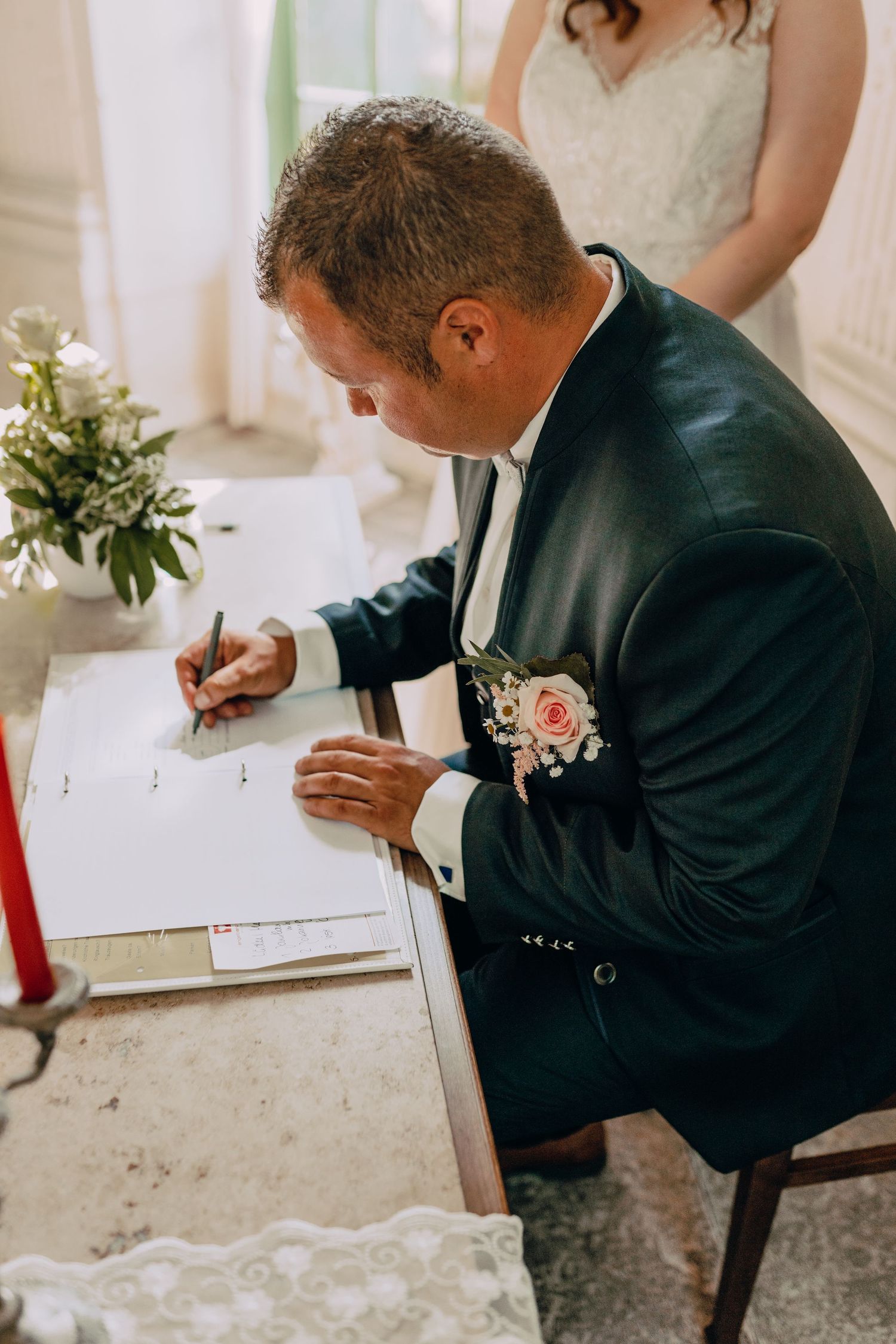 Groom in navy suit with pink boutonniere signs marriage certificate at wedding ceremony while bride stands nearby.