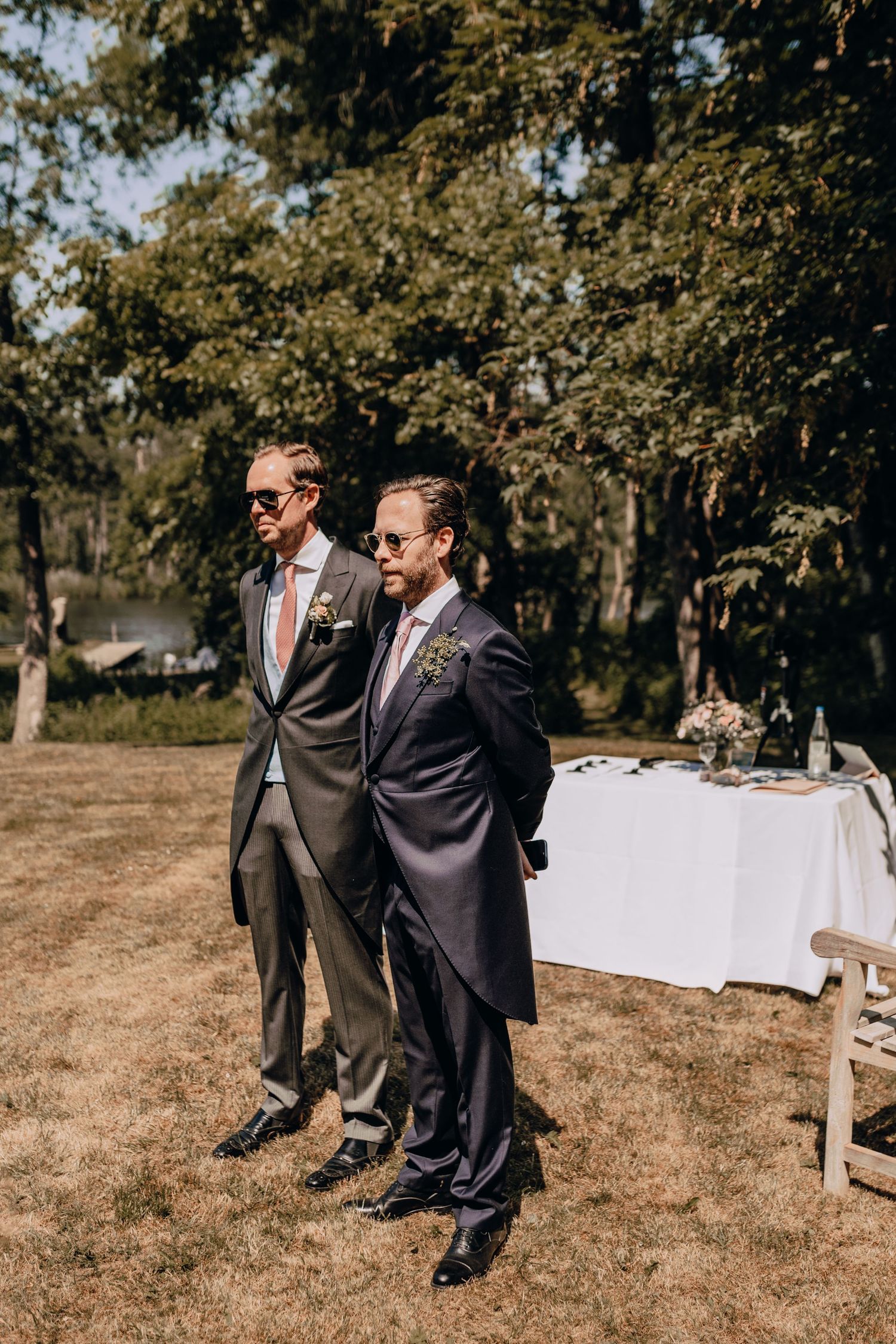 Two men in formal suits stand on lawn at outdoor wedding reception with white table in background.