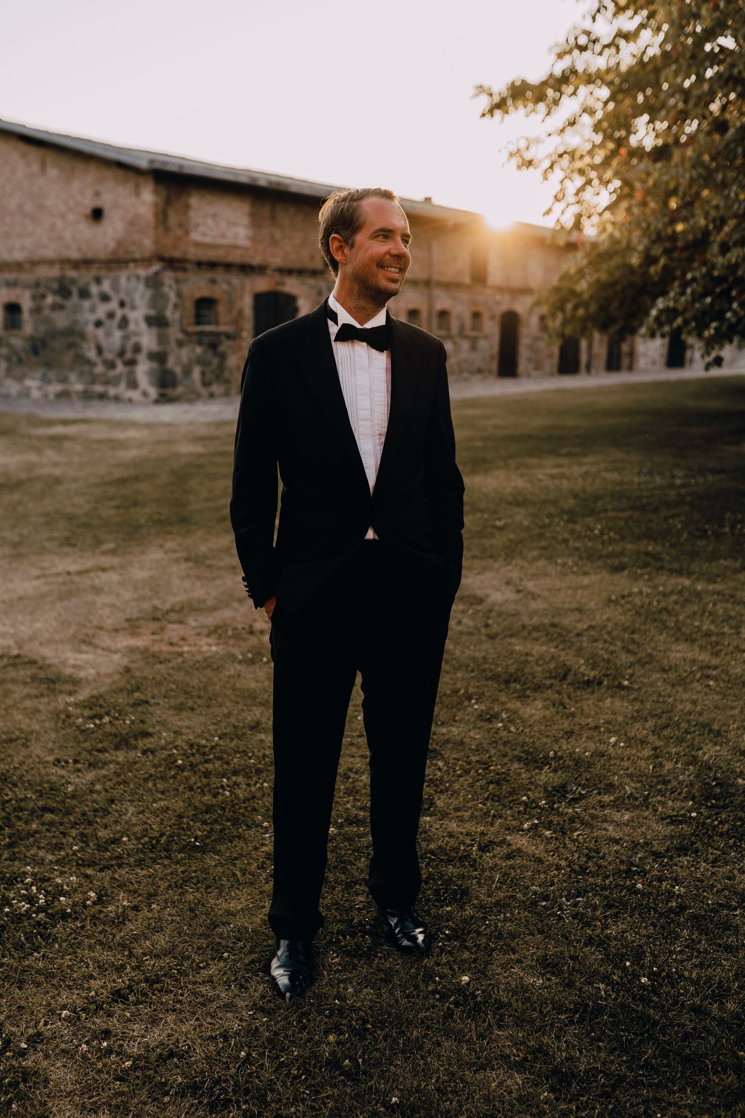Groom in classic black tuxedo standing on lawn at sunset near stone building.