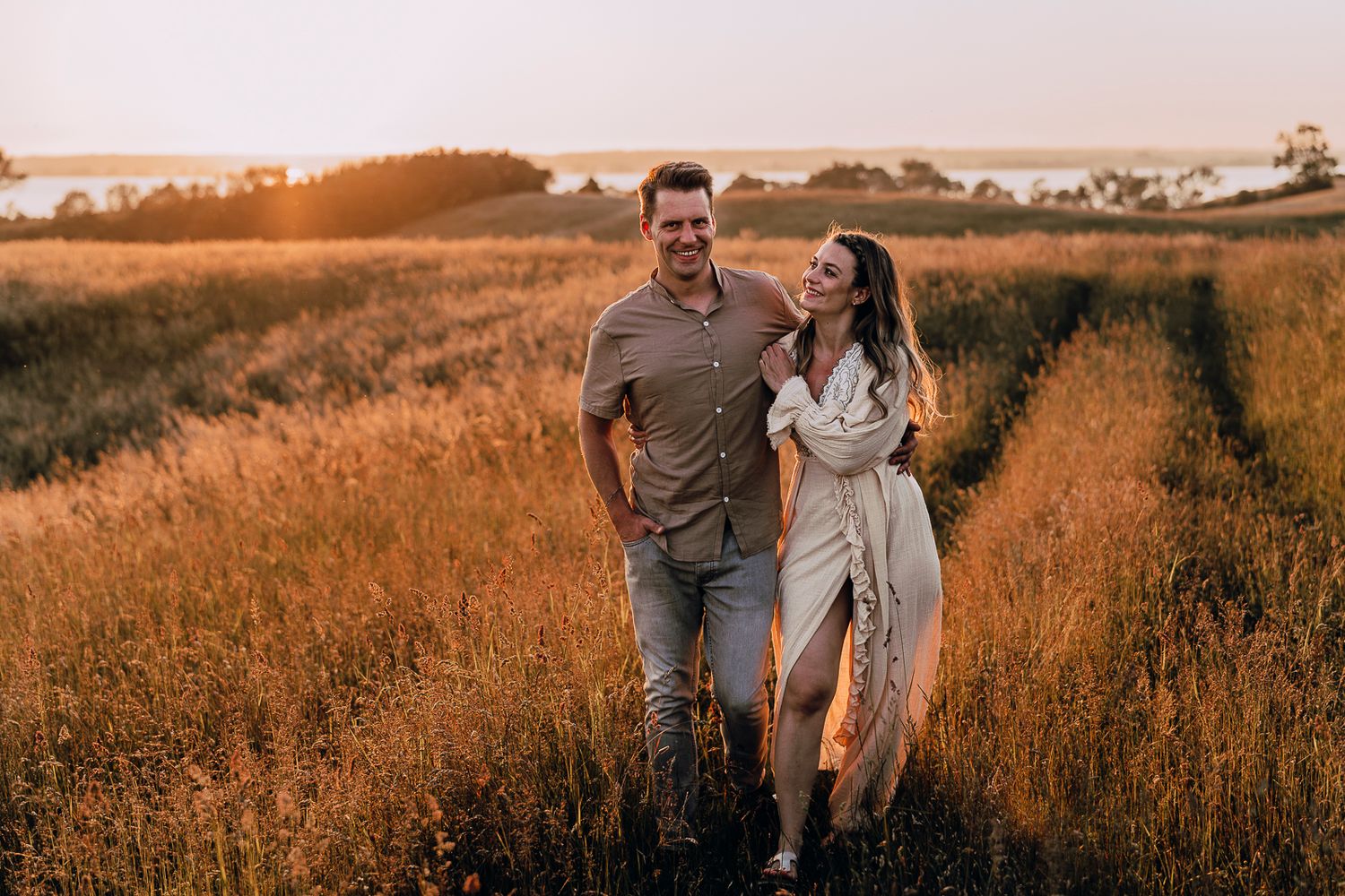 Romantic couple holding hands while walking through golden grass field at sunset, creating silhouettes against warm sky.