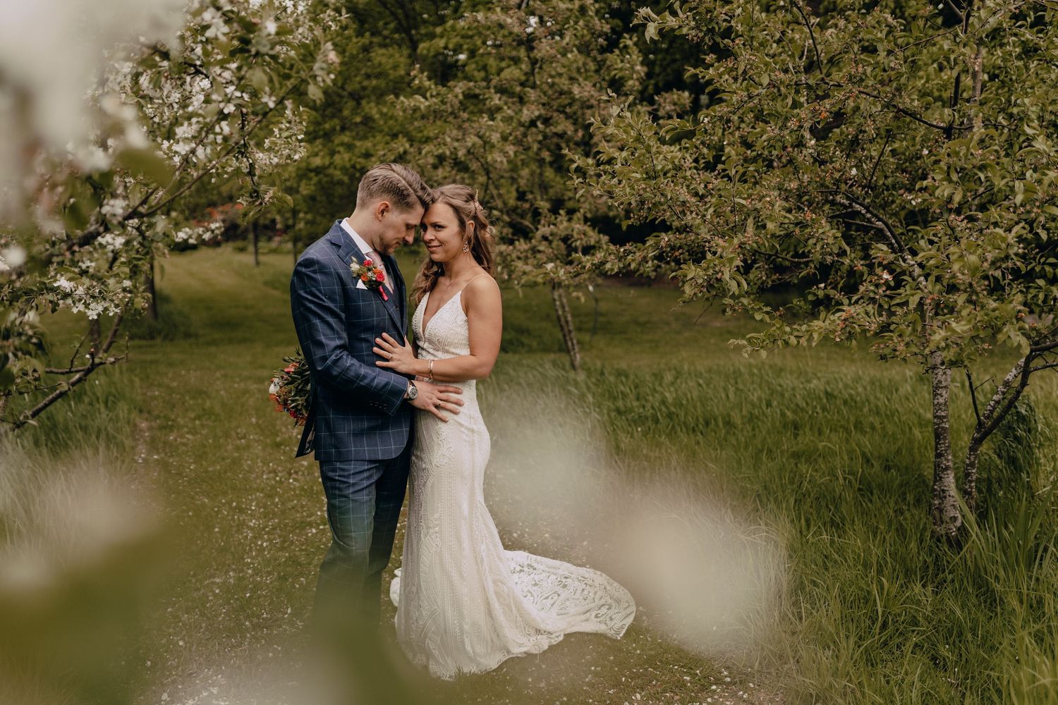 Romantic wedding portrait of couple embracing in an orchard with soft focus foreground elements.